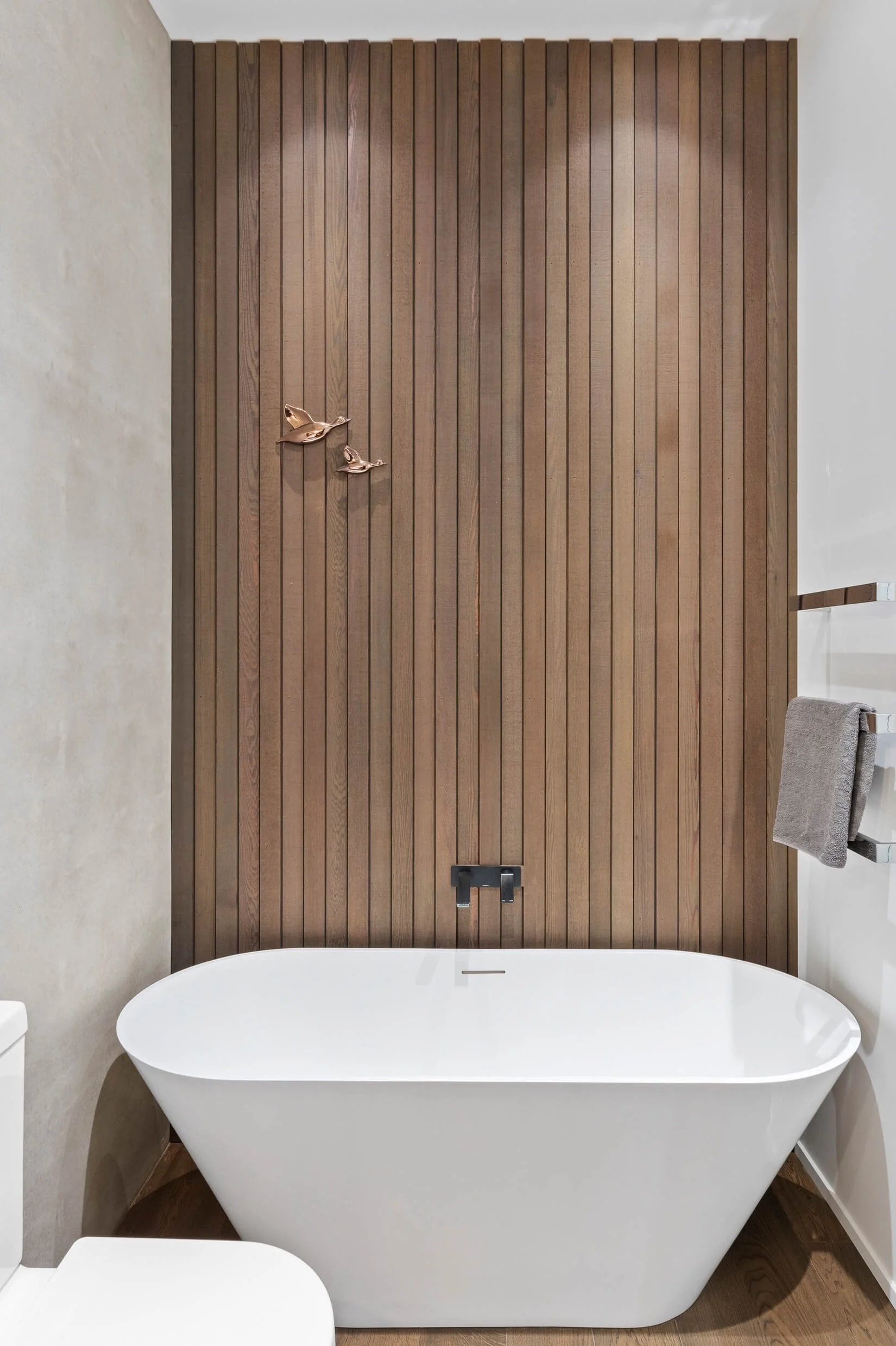 Freestanding white bathtub in a bathroom with a tall wooden paneled wall behind it, decorated with a bronze bird sculpture, with a towel hanging on a rack to the right.