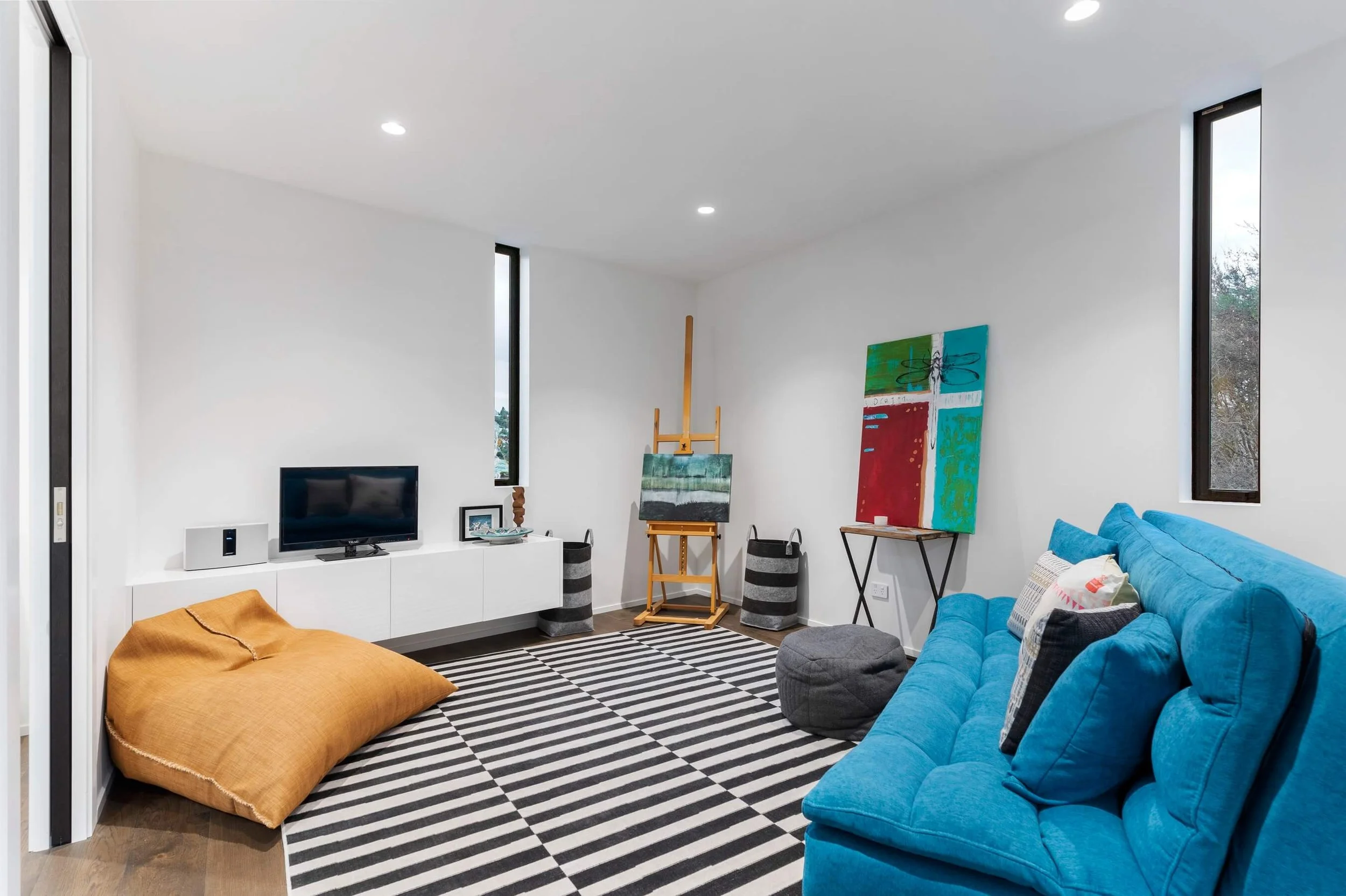 Modern living room with white walls and high ceiling, featuring a blue sofa, orange bean bag, black and white striped rug, wall-mounted TV, colorful abstract paintings, and floor-to-ceiling windows.