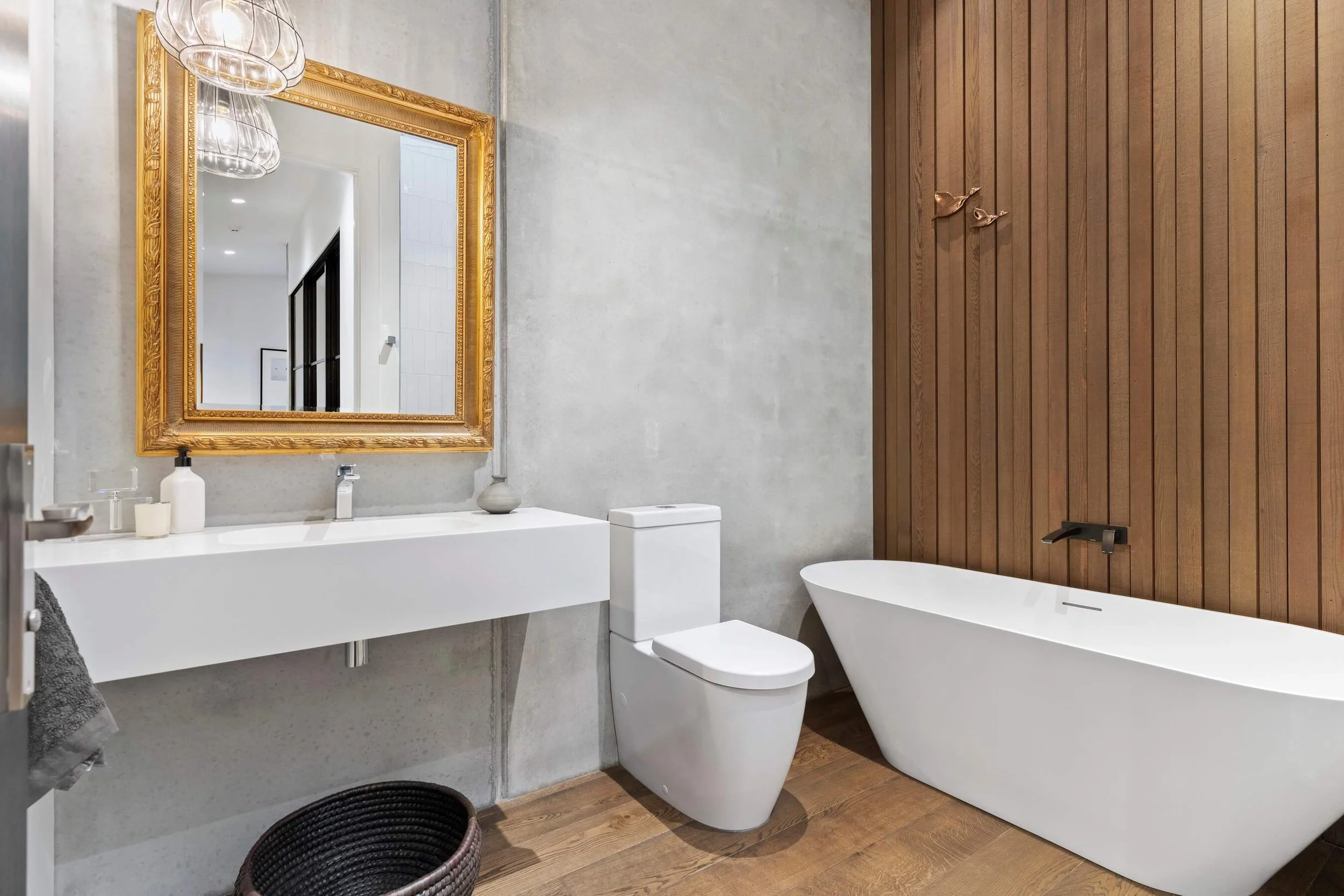 Modern bathroom with a large gold-framed mirror, a white floating sink with toiletries, a white toilet, a freestanding bathtub, wood paneling on one wall, and decorative bird sculptures on the wall.