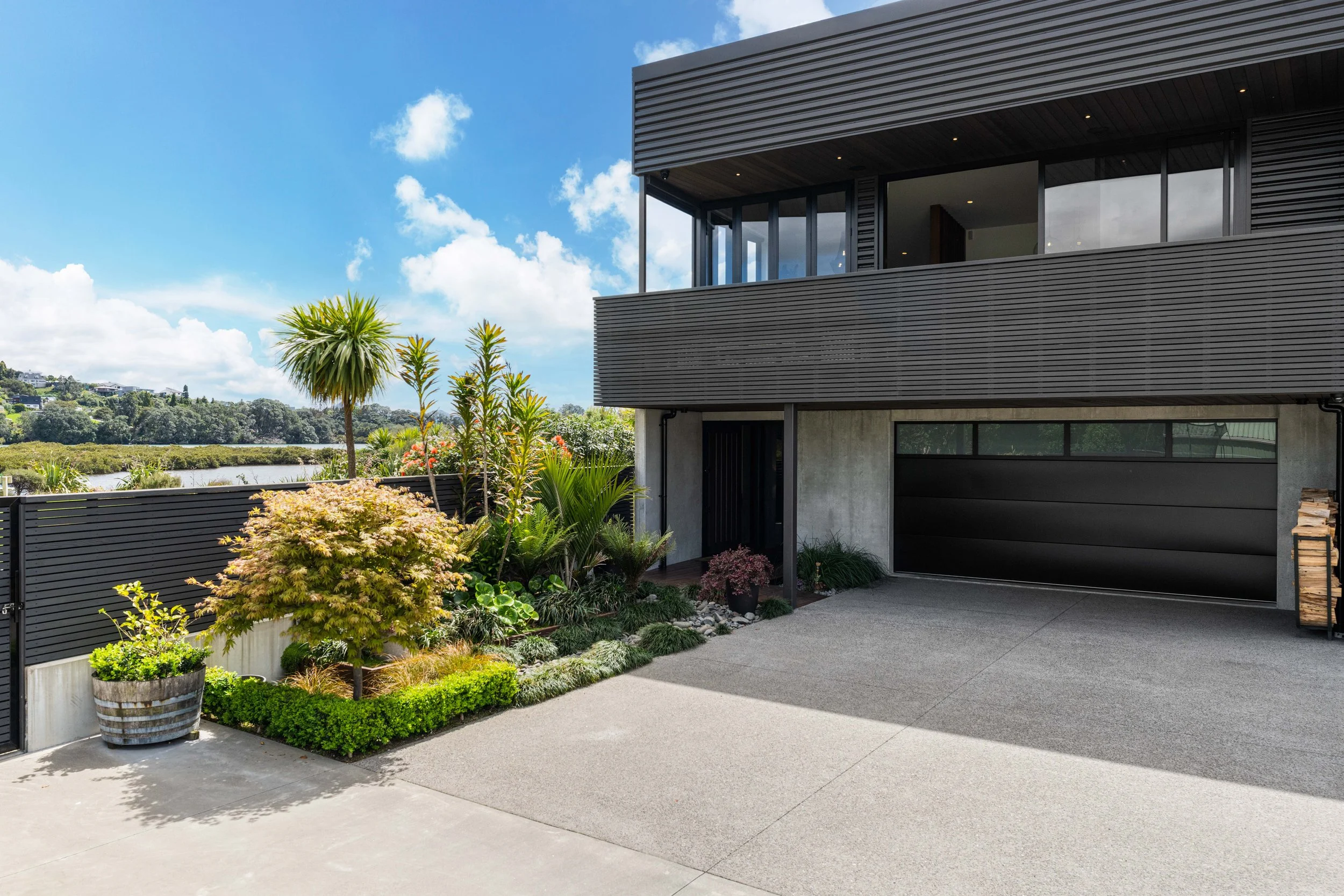 Modern house with black siding, large glass windows, and a driveway, surrounded by landscaped garden with various plants and trees under a blue sky with clouds.
