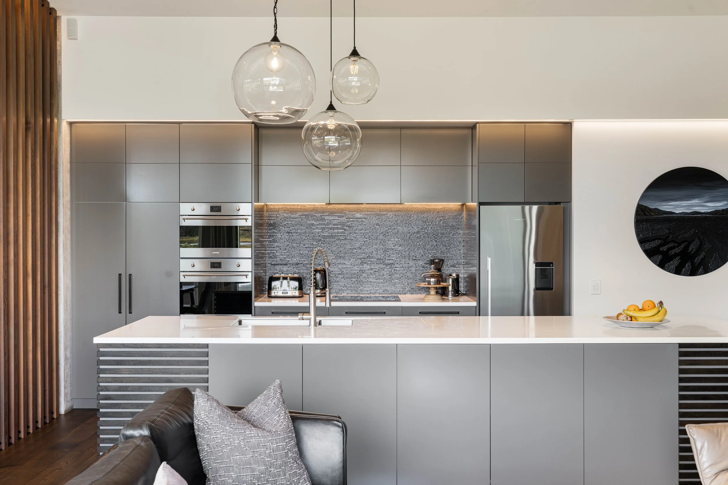 Modern kitchen with gray cabinets, a white island, glass pendant lights, stainless steel appliances, a textured backsplash, and a bowl of bananas on the counter.
