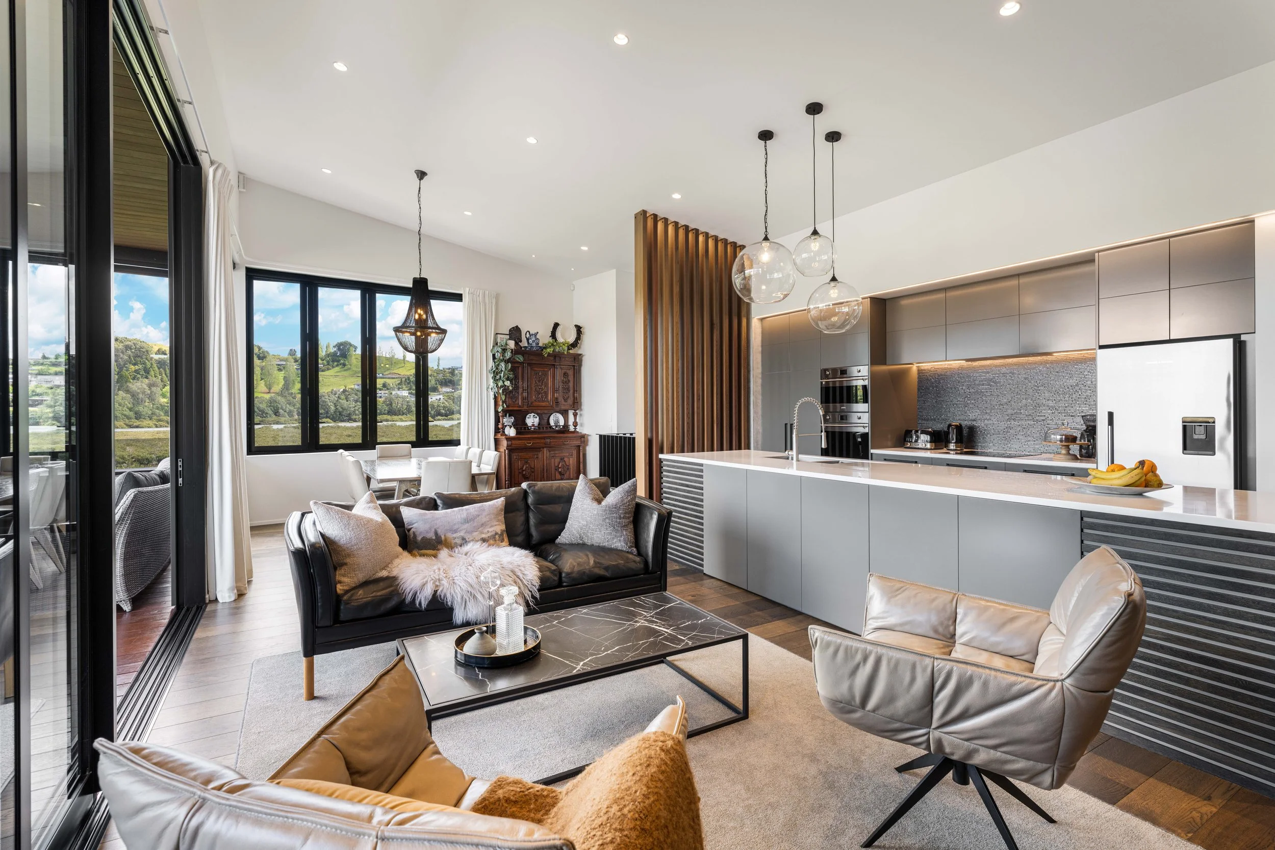 Open-concept living room and kitchen with large windows revealing a green landscape, modern furniture, and pendant lights over the kitchen island.