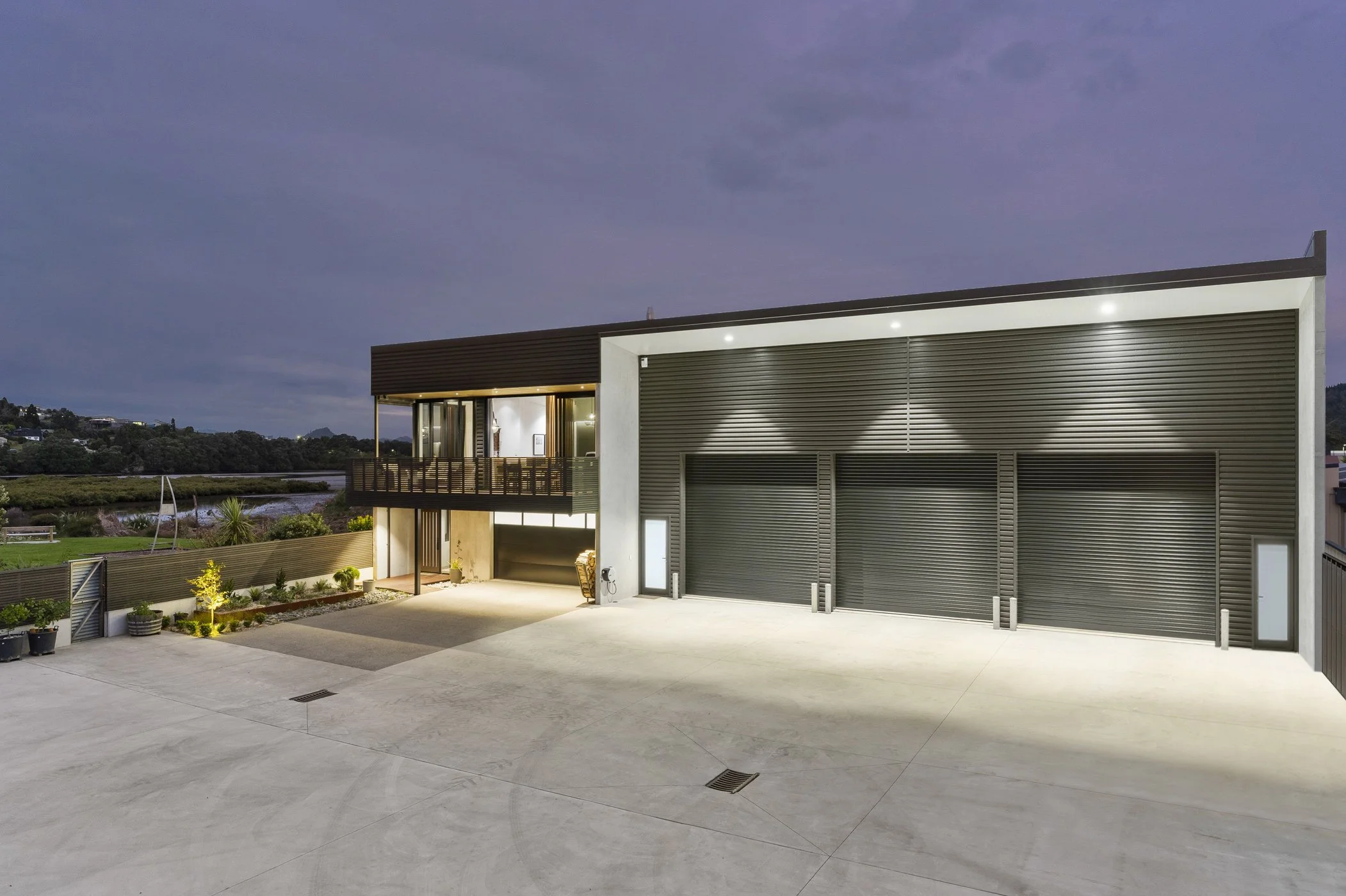 Modern house exterior at twilight with garage doors, balcony, outdoor lighting, and landscaped yard.