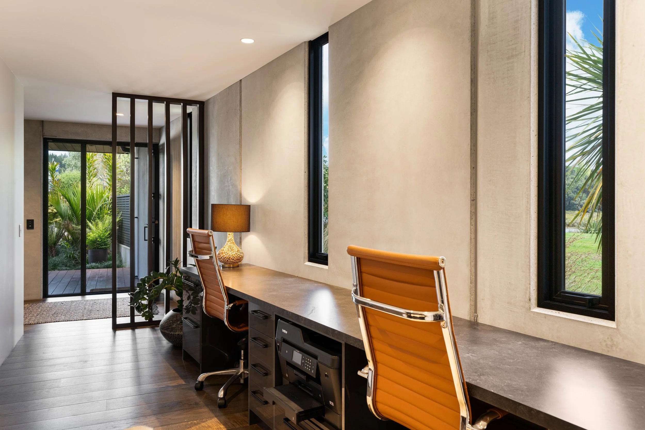 Modern office space with a long desk, orange office chairs, a table lamp, large windows showing outdoor greenery, and a sliding glass door leading outside.
