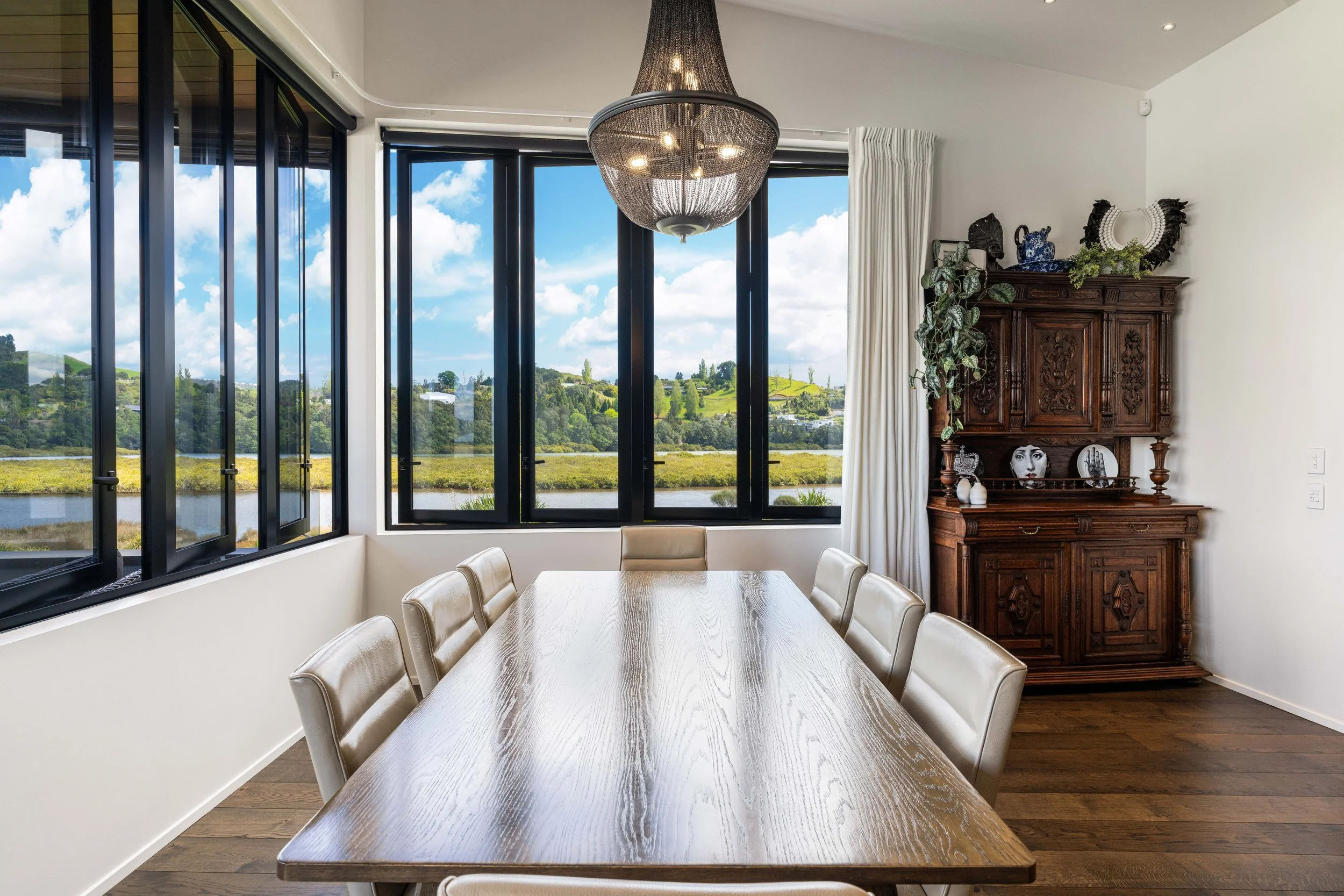 A dining room with a wooden table surrounded by cream-colored chairs, large windows showing a scenic view of the Moana Anu estuary and hills, a decorative dark wood cabinet with plants and decorative items, and a chandelier hanging from the ceiling.