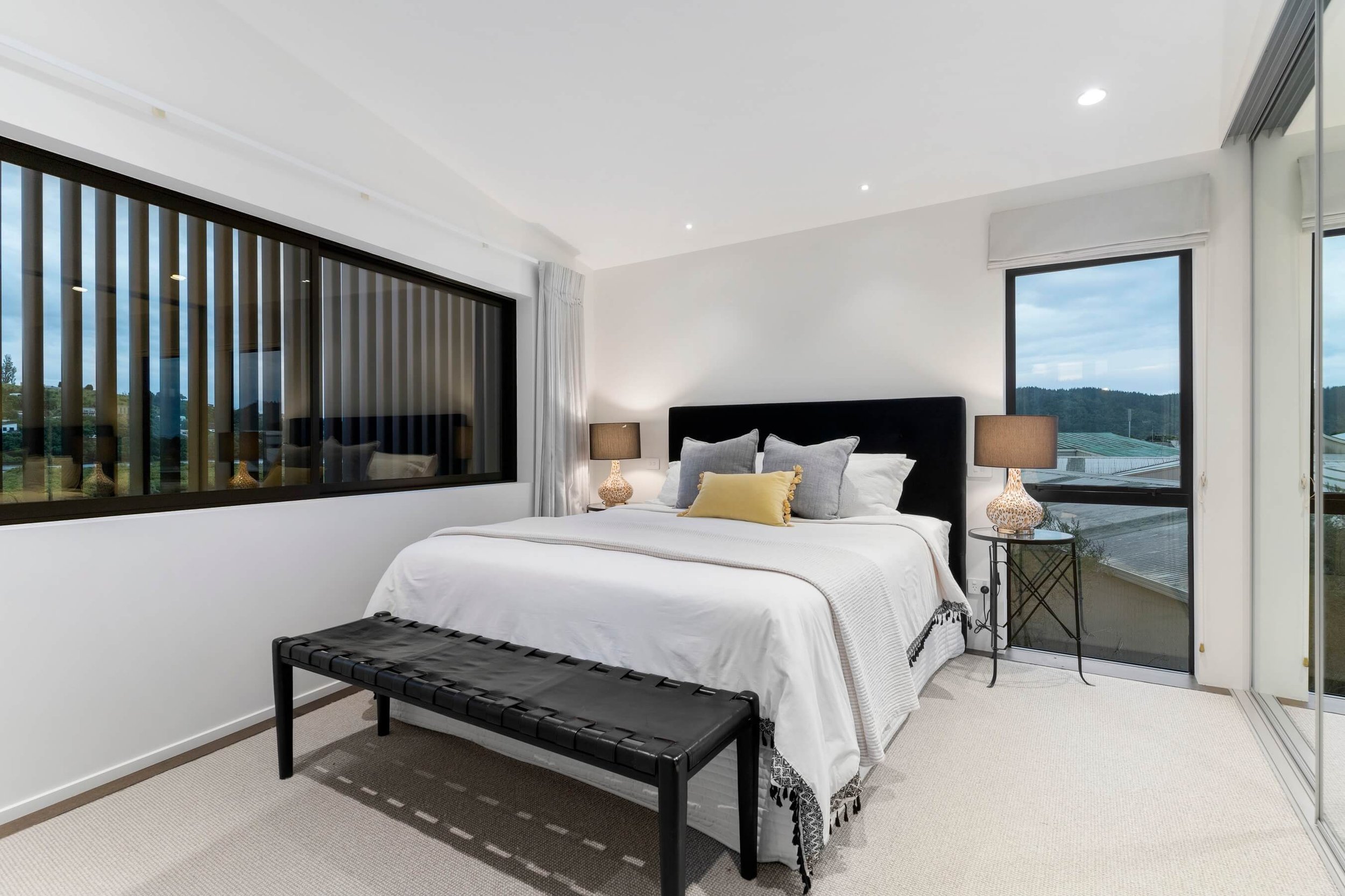 A modern bedroom with a large bed topped with white bedding and multiple pillows, black headboard, bedside tables with lamps, large windows showing a scenic outdoor view, and a sliding glass door on the right.