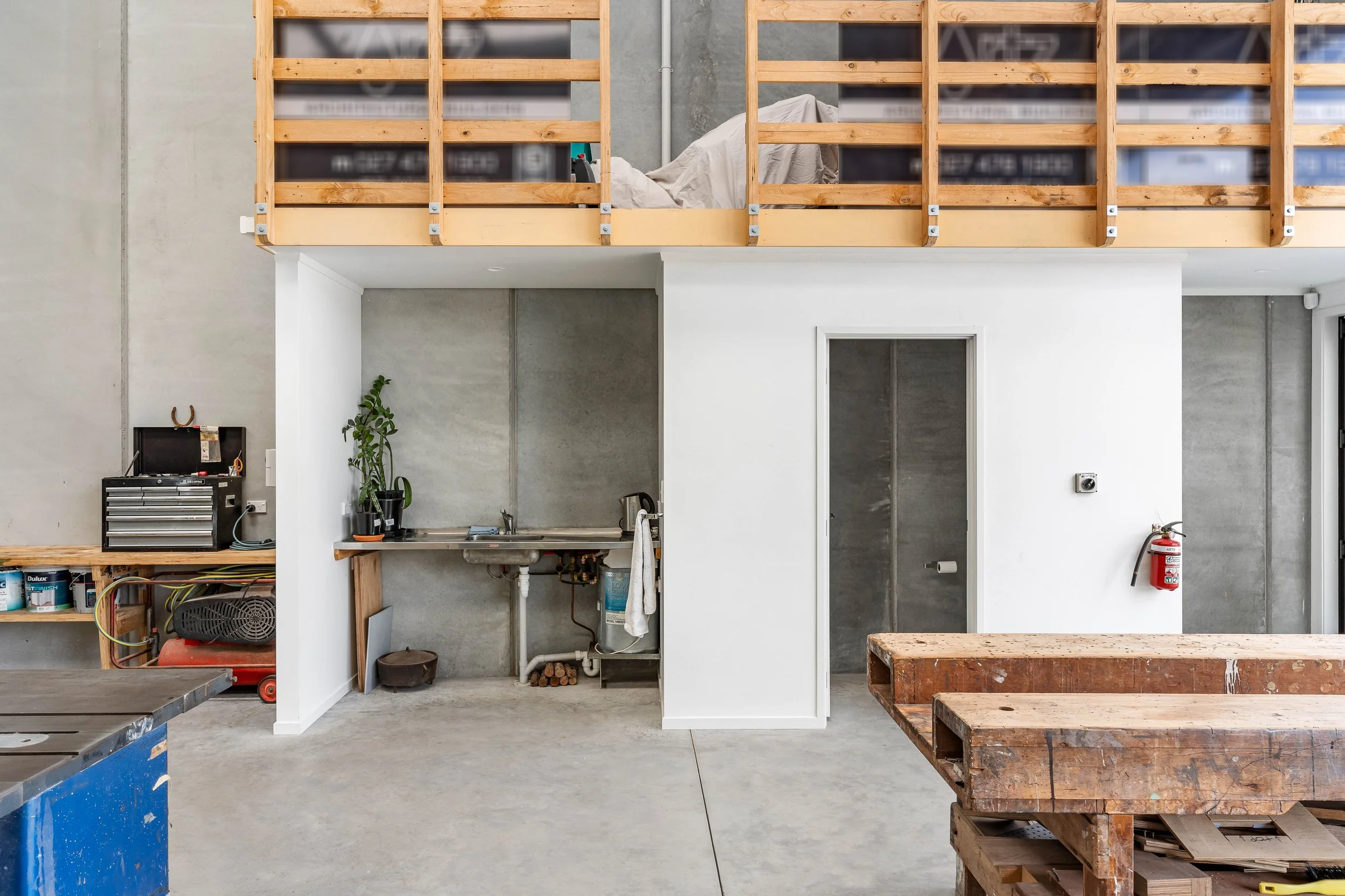 Interior of a building under construction with exposed concrete walls, a wooden loft structure, a small kitchenette with a sink, a plant, a fire extinguisher, and wooden pallets in the foreground.