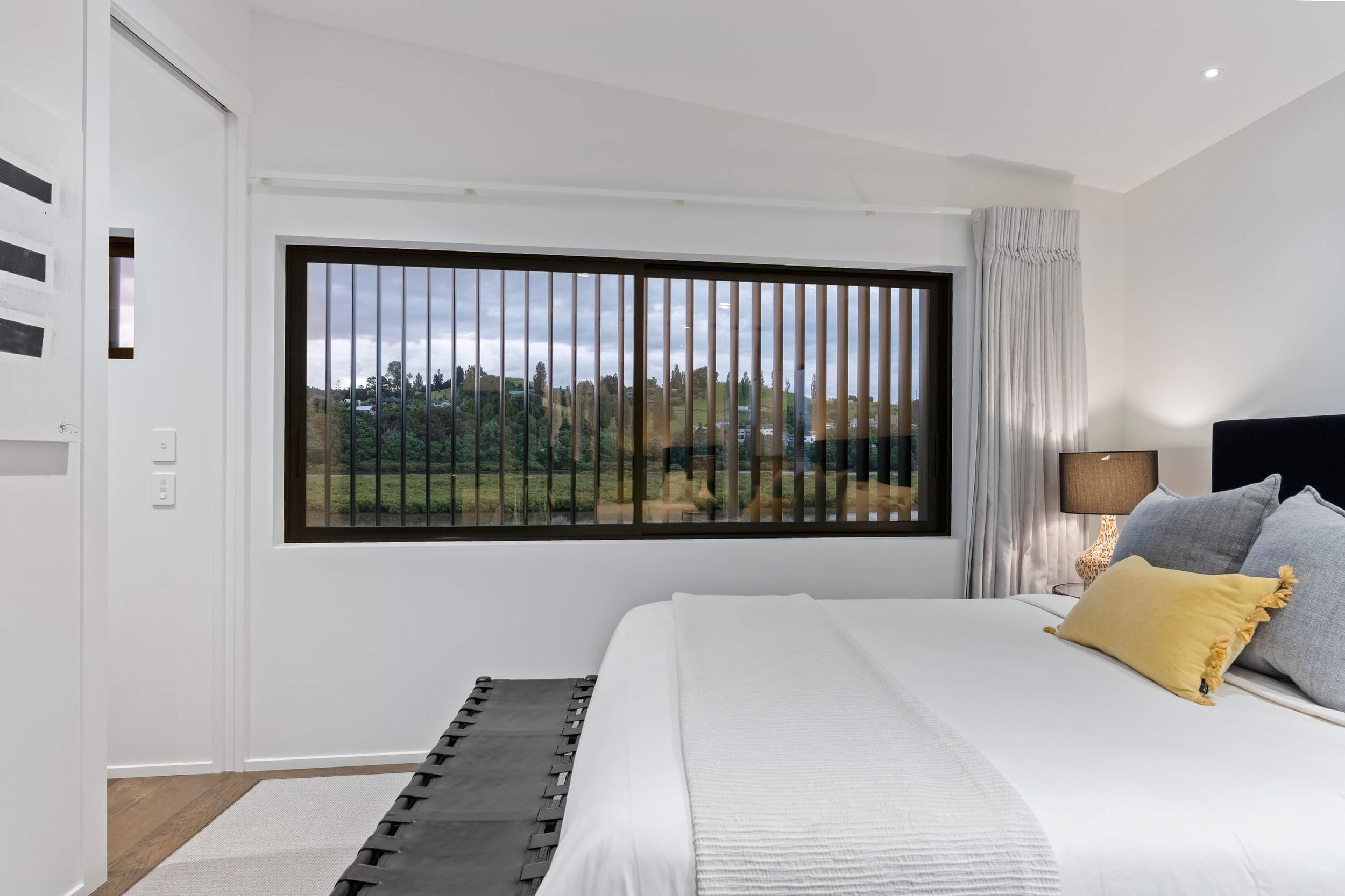 A bedroom with a large window overlooking a green landscape and cloudy sky. The room has white walls, a bed with gray and yellow pillows, a side table with a lamp, and white curtains.