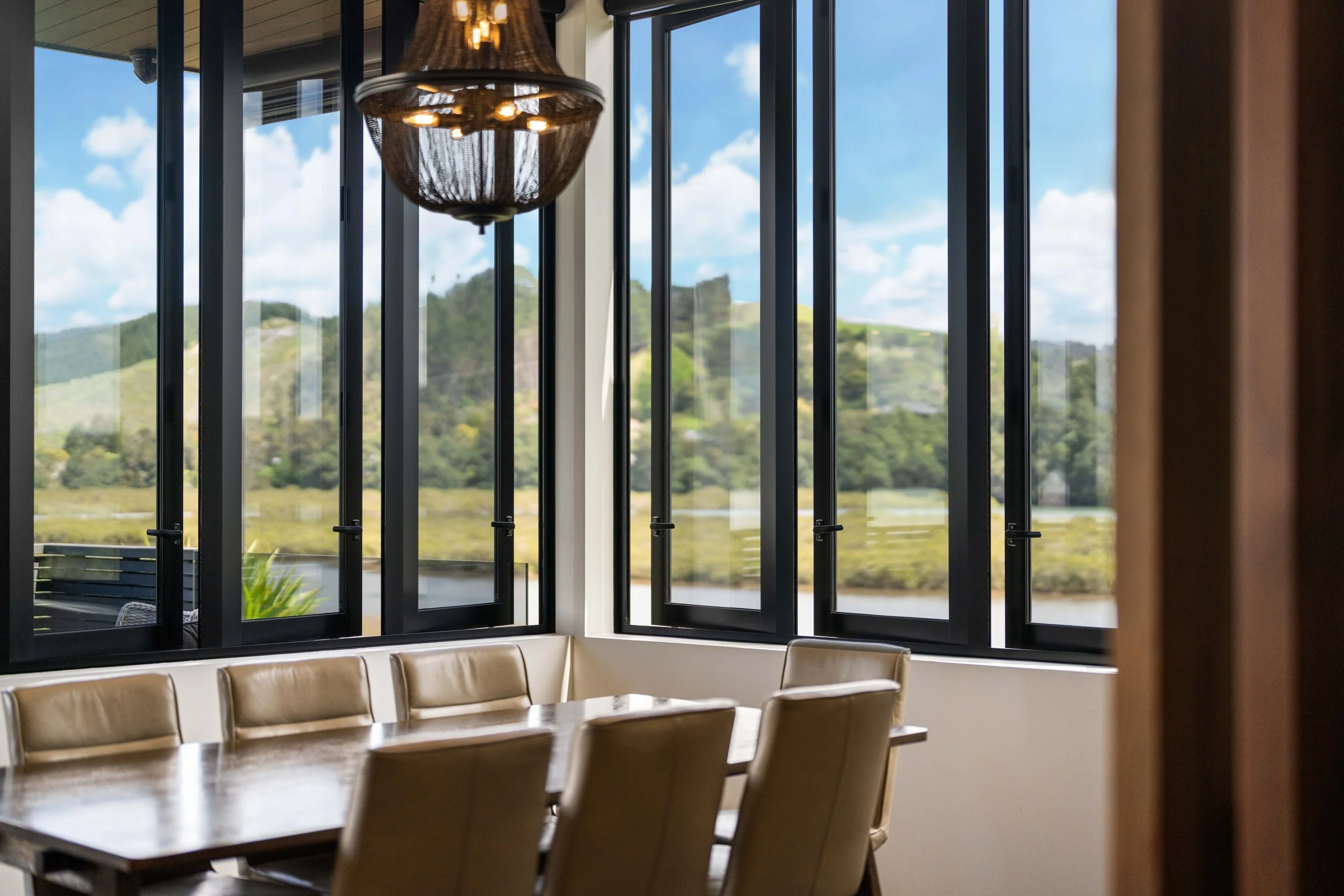 A dining room table surrounded by beige chairs with leather upholstery, large open windows showing a bright blue sky with clouds and a scenic landscape with green hills.