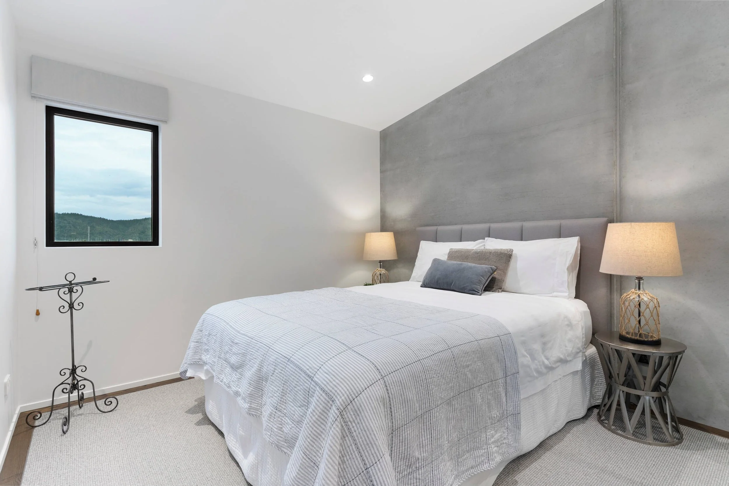 A modern bedroom with a bed that has white and gray striped bedding, a gray upholstered headboard, and decorative pillows. There are two matching beige lamps on side tables, a window showing a scenic view of green hills and cloudy sky, and a decorati