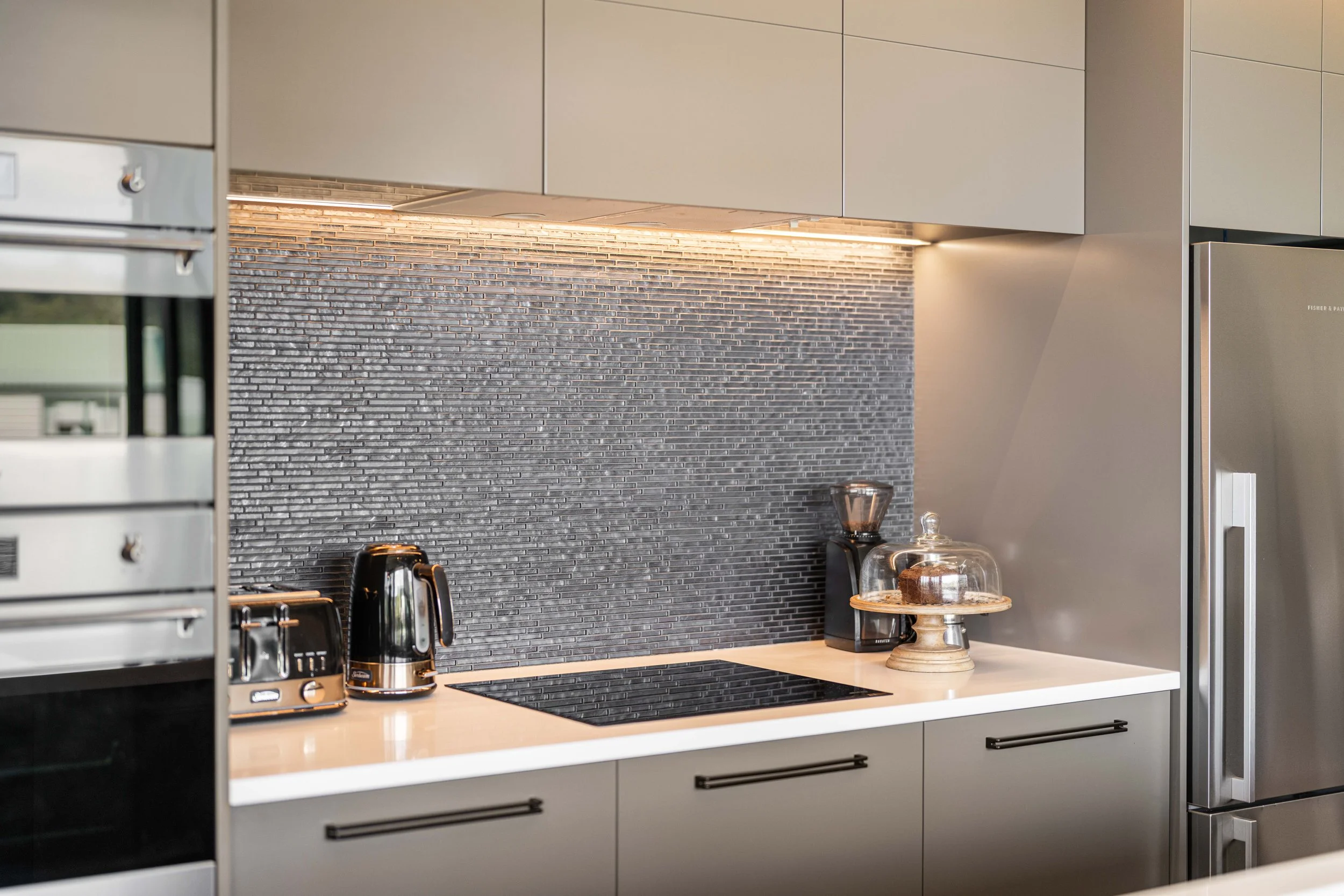 Modern kitchen with gray cabinets, black textured backsplash, stainless steel appliances, black toaster and kettle, clear glass cake stand with cake, and breakfast pastries on a wooden cake stand.