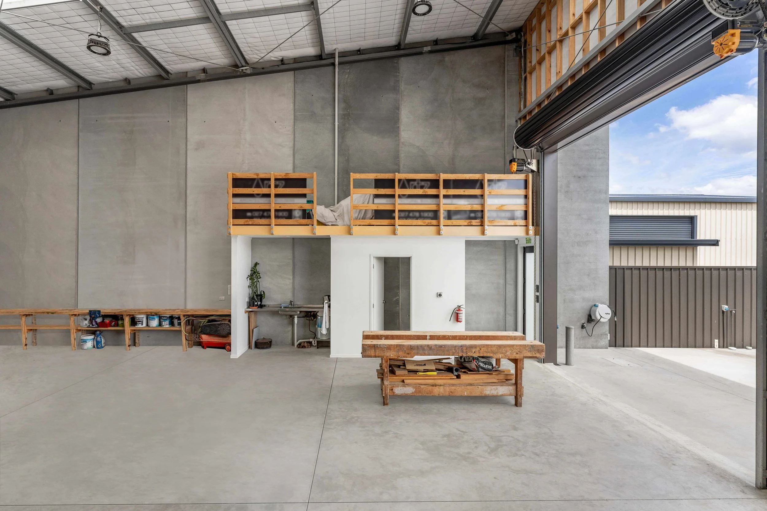 Interior view of an industrial space with a high ceiling, and a partially open garage door showing a blue sky with clouds. There are shelves with paint cans, a small kitchen area, and wooden structures.