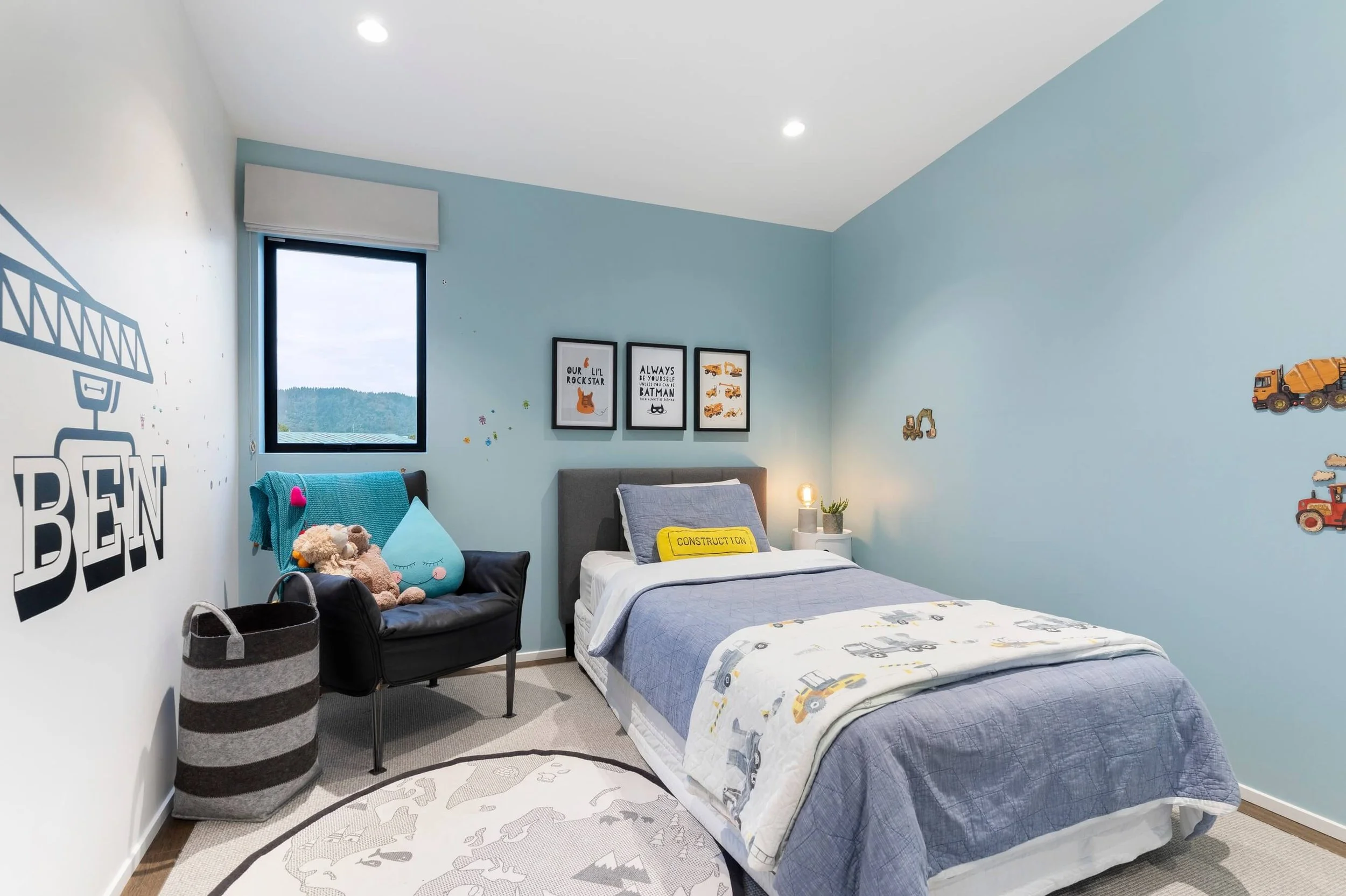 A bedroom with light blue walls, a window showing an outdoor view, a twin bed with a construction-themed blanket, a gray headboard, a small white nightstand with a lamp and plants, framed art on the wall featuring construction vehicles and quotes, a 