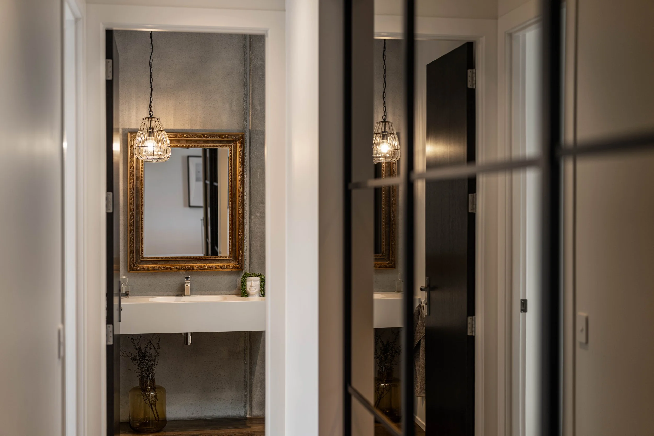 A hallway view into a bathroom, with a mirror reflecting a hanging light fixture, a small vanity with a soap dispenser and a decorative plant, and another mirror in the distance.