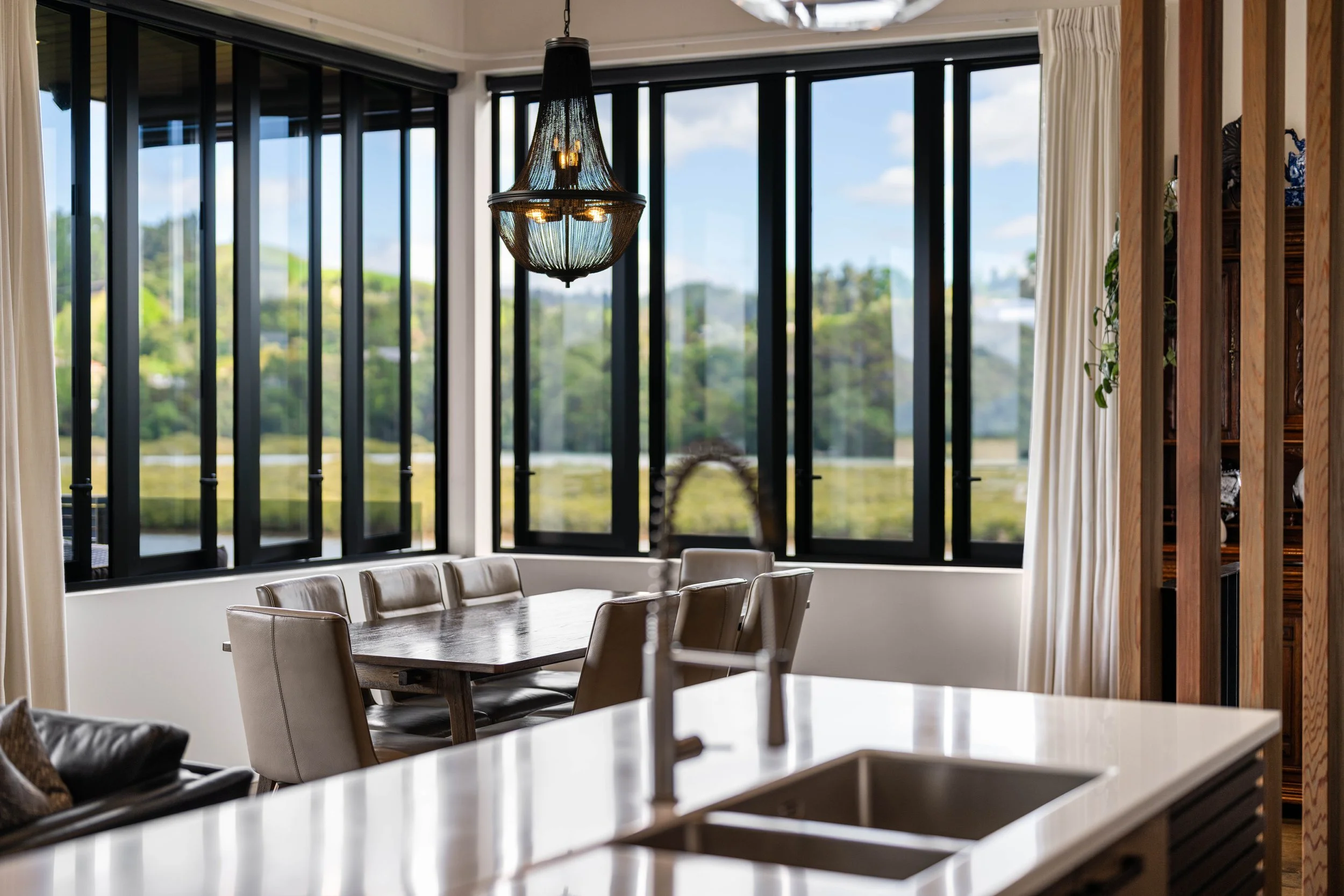 A dining room with large windows overlooking greenery, a wooden dining table surrounded by beige chairs, a black pendant chandelier hanging from the ceiling, and a wooden and glass bookshelf on the right side.