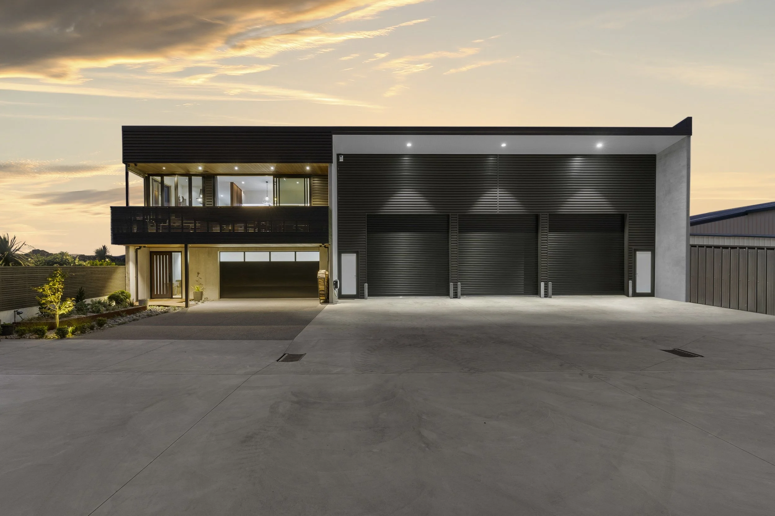 Modern house with black metal garage doors and a balcony, illuminated at dusk with the sky showing sunset colors.