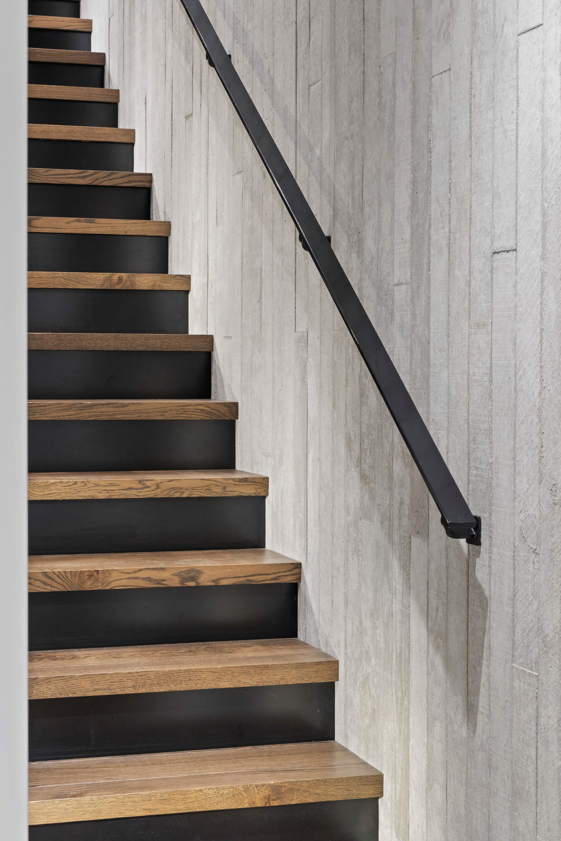 Photo showing a modern staircase with wooden steps, a black riser, and a black metal railing, against a concrete wall.