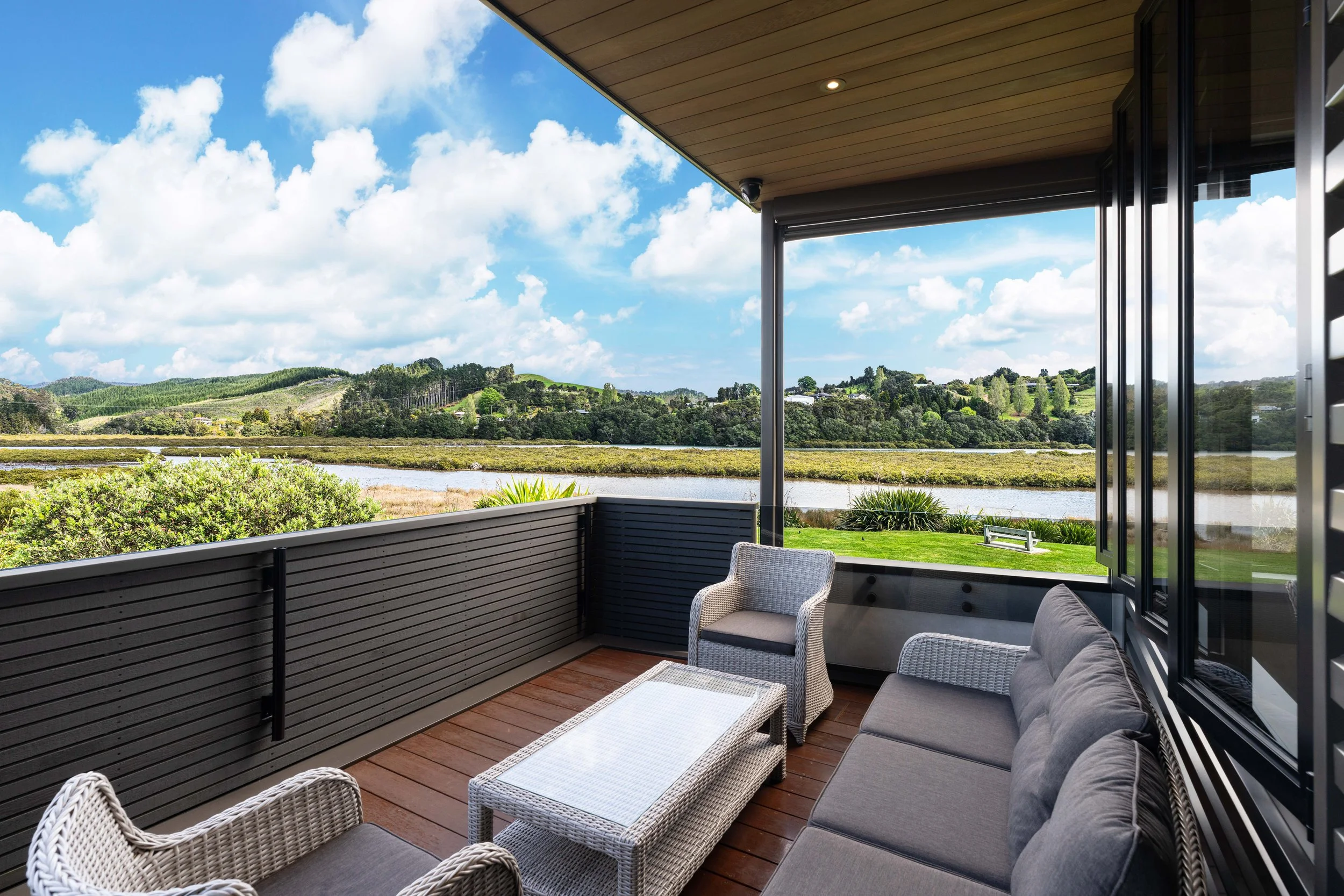 Balcony with outdoor furniture overlooking a river, trees, and hills under blue sky with clouds.