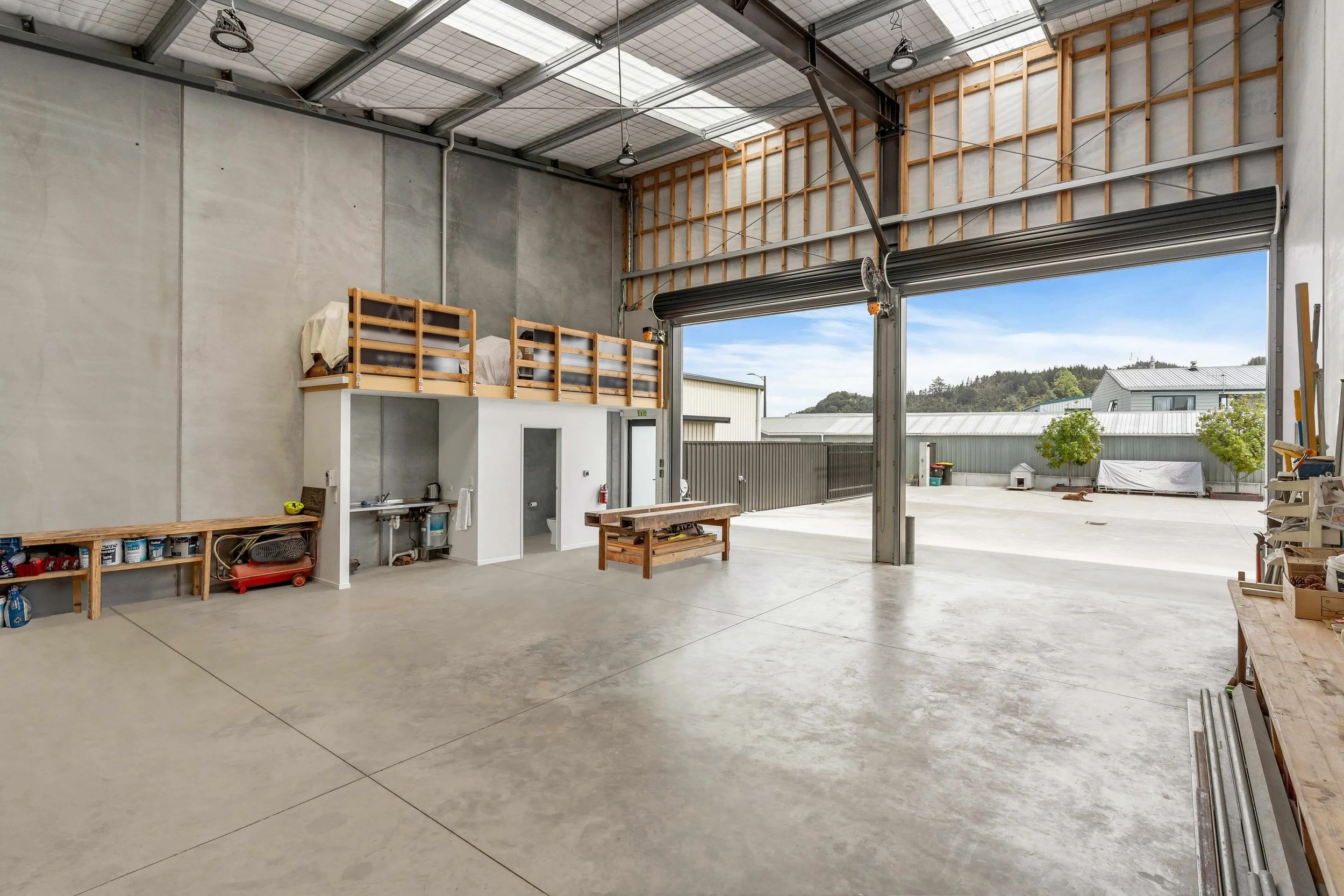 Interior of an industrial warehouse with high ceiling, concrete floors, and large open doors showing an outdoor view with a clear sky, buildings, trees, and a dog lying on the ground.