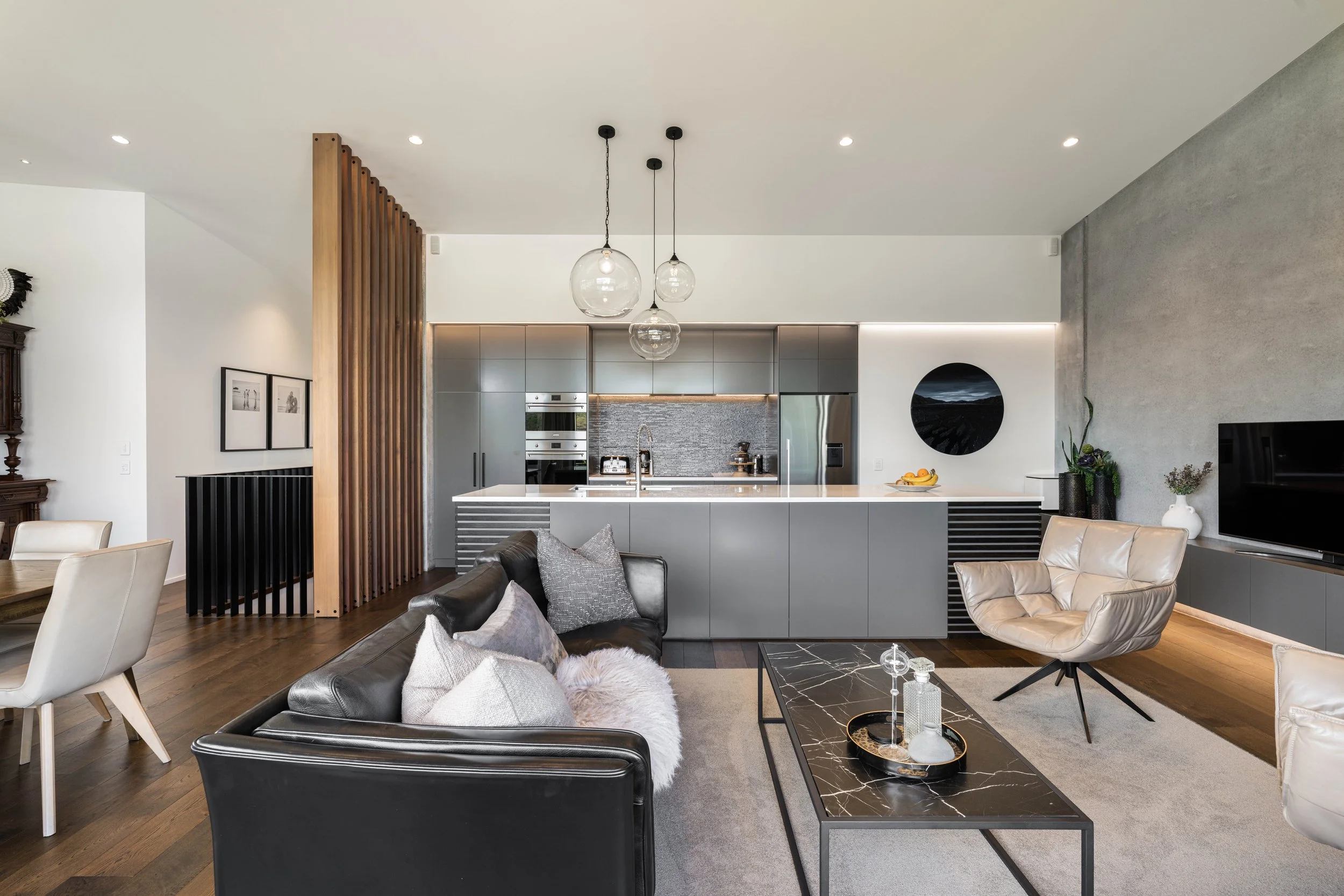 Modern open-concept living room and kitchen with neutral decor, large glass pendant lights, and a mix of furniture including a black leather sofa and beige armchair.