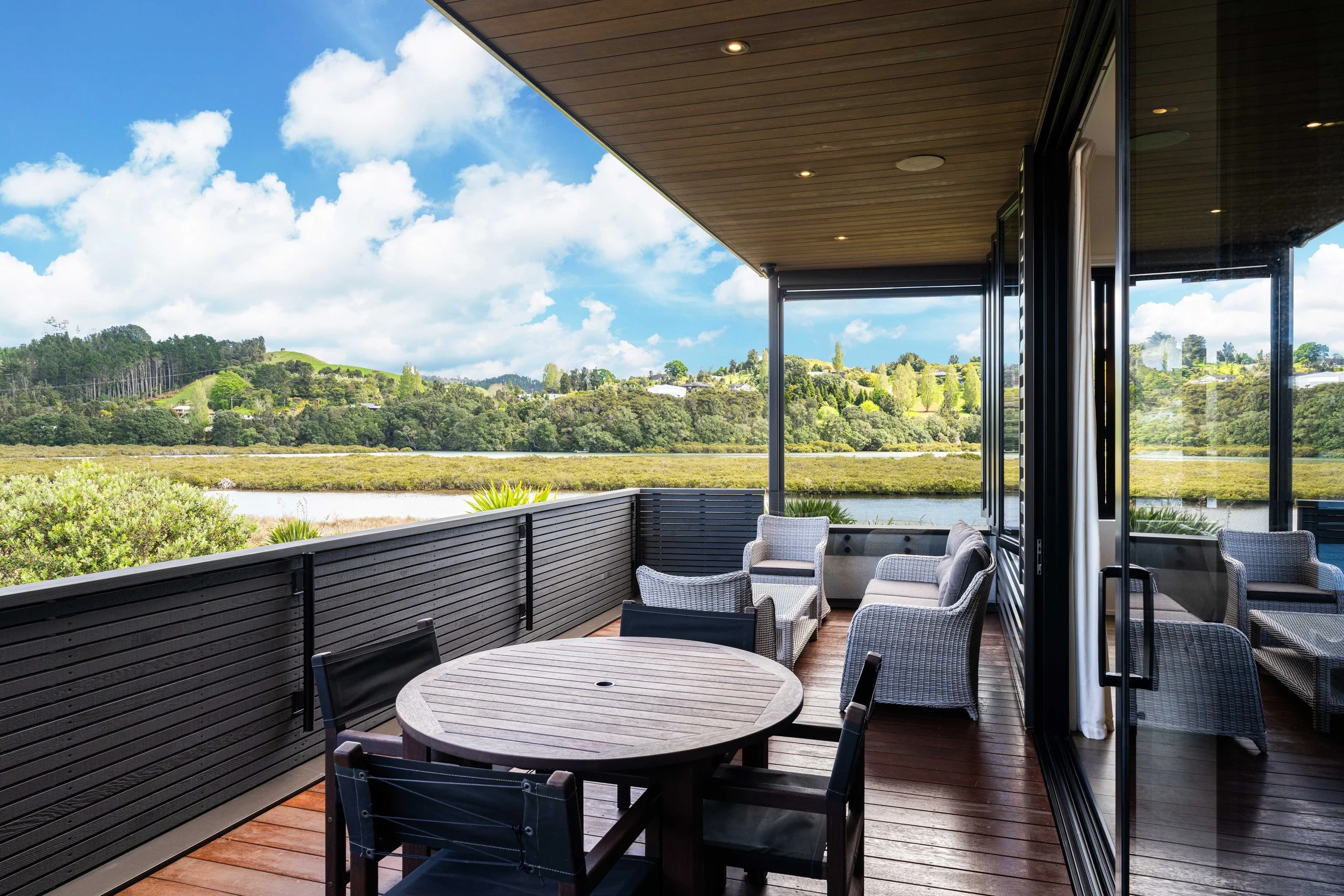 Outdoor balcony with a round table and chairs, overlooking a lush green landscape with trees, hills, and a partly cloudy sky.