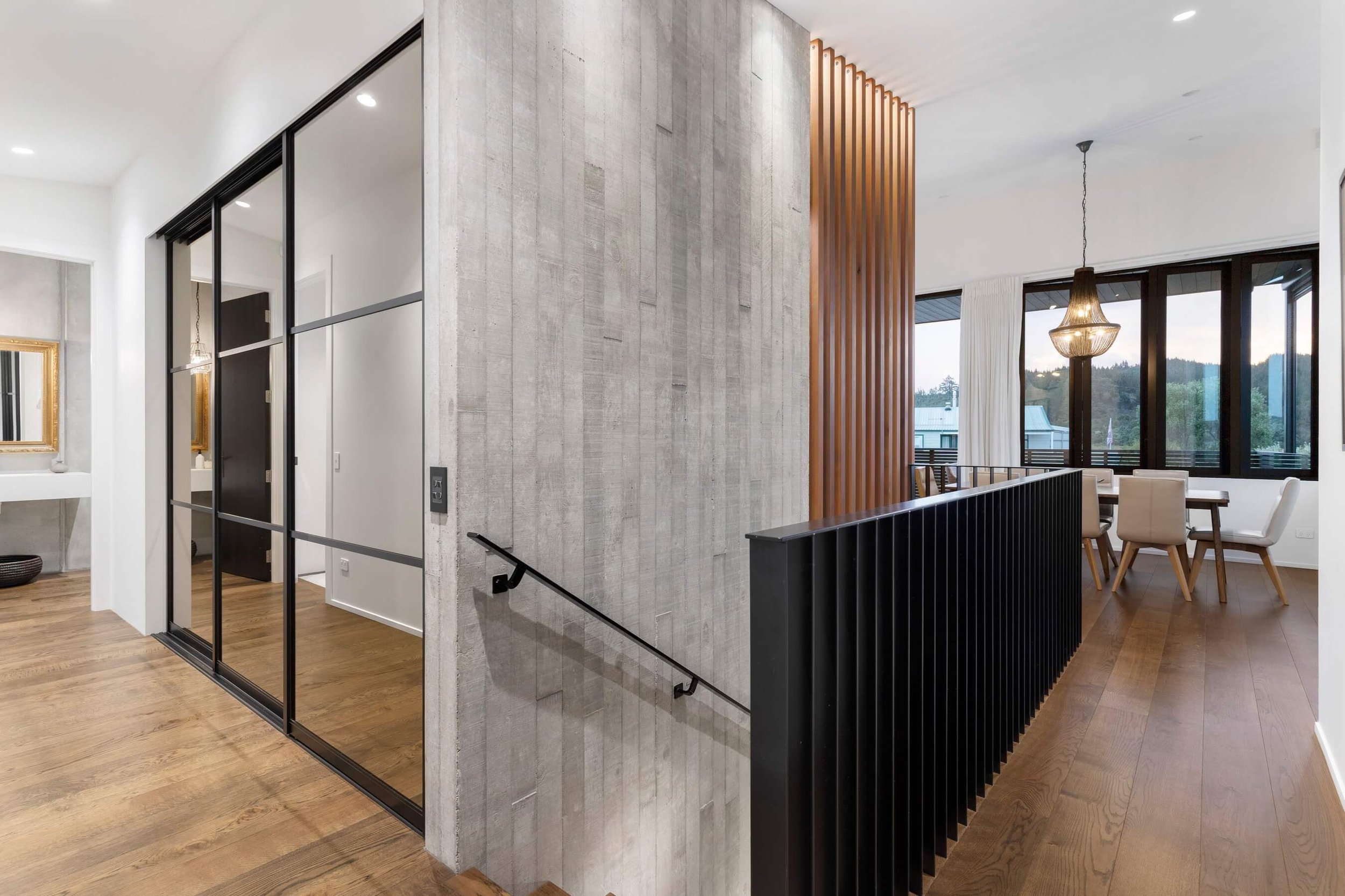 Modern interior with a concrete and wooden partition, black metal-framed glass sliding doors, wooden flooring, and a dining area with large windows, a chandelier, and white and wooden furniture.