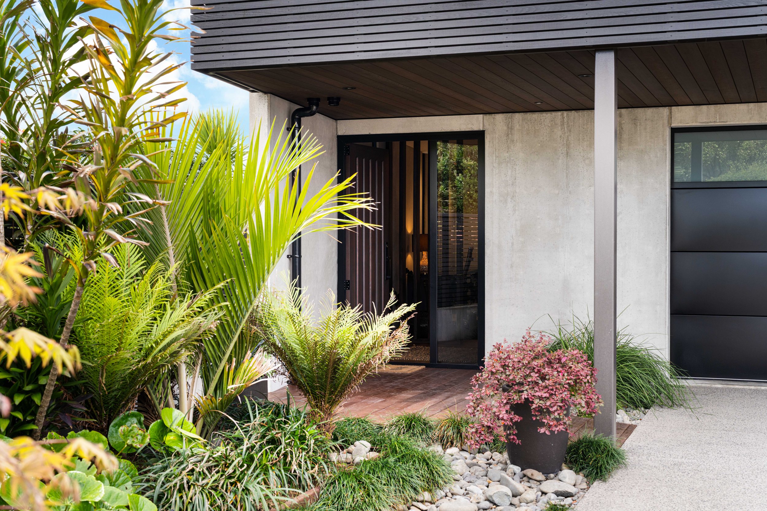 The image shows a modern house entrance with concrete walls, and a cedar and glass front door. There is greenery in the foreground, including various plants, bushes, and flowers, along with a small landscape of rocks and a concrete pathway.