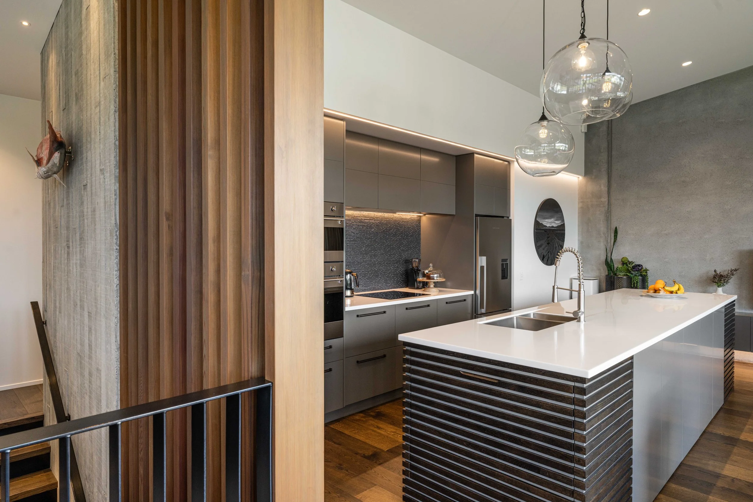 Modern kitchen with gray cabinetry, a white kitchen island with dark wood accents, and pendant lights with glass globes hanging from the ceiling. There is a wooden slat partition on the left side of the image and a decorative wall hanging on the left