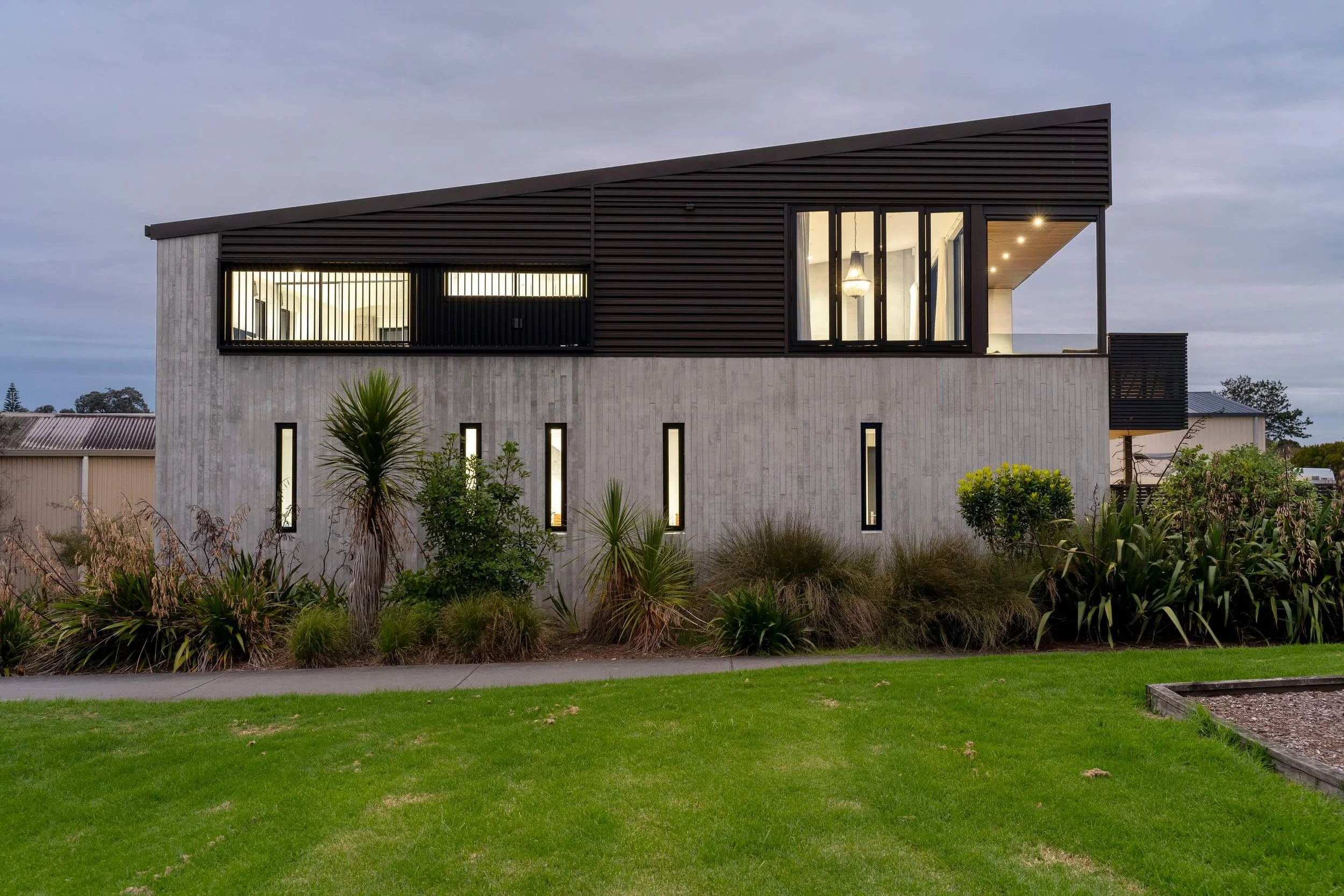 Modern two-story house with a concrete and dark siding exterior, large windows, and a garden with various plants and grass in the foreground.