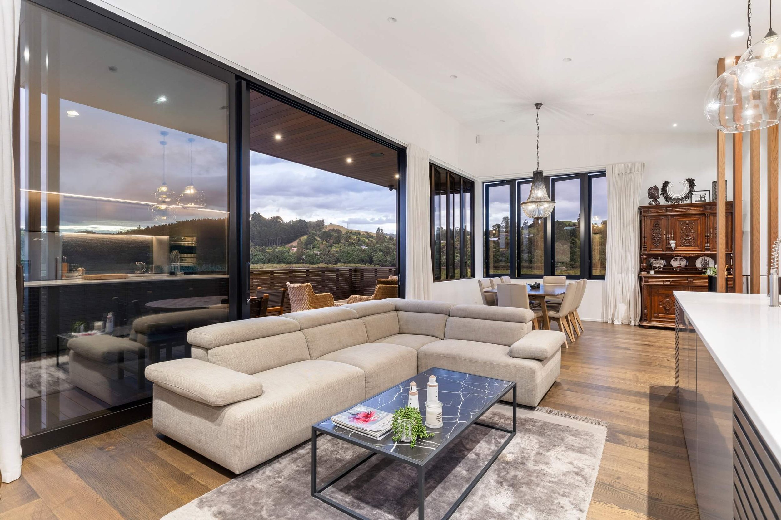 Modern open-concept living room with large windows and views of greenery and hills, featuring a beige sectional sofa, black coffee table with magazines and plants, wooden flooring, and a wooden cabinet with decorative items.