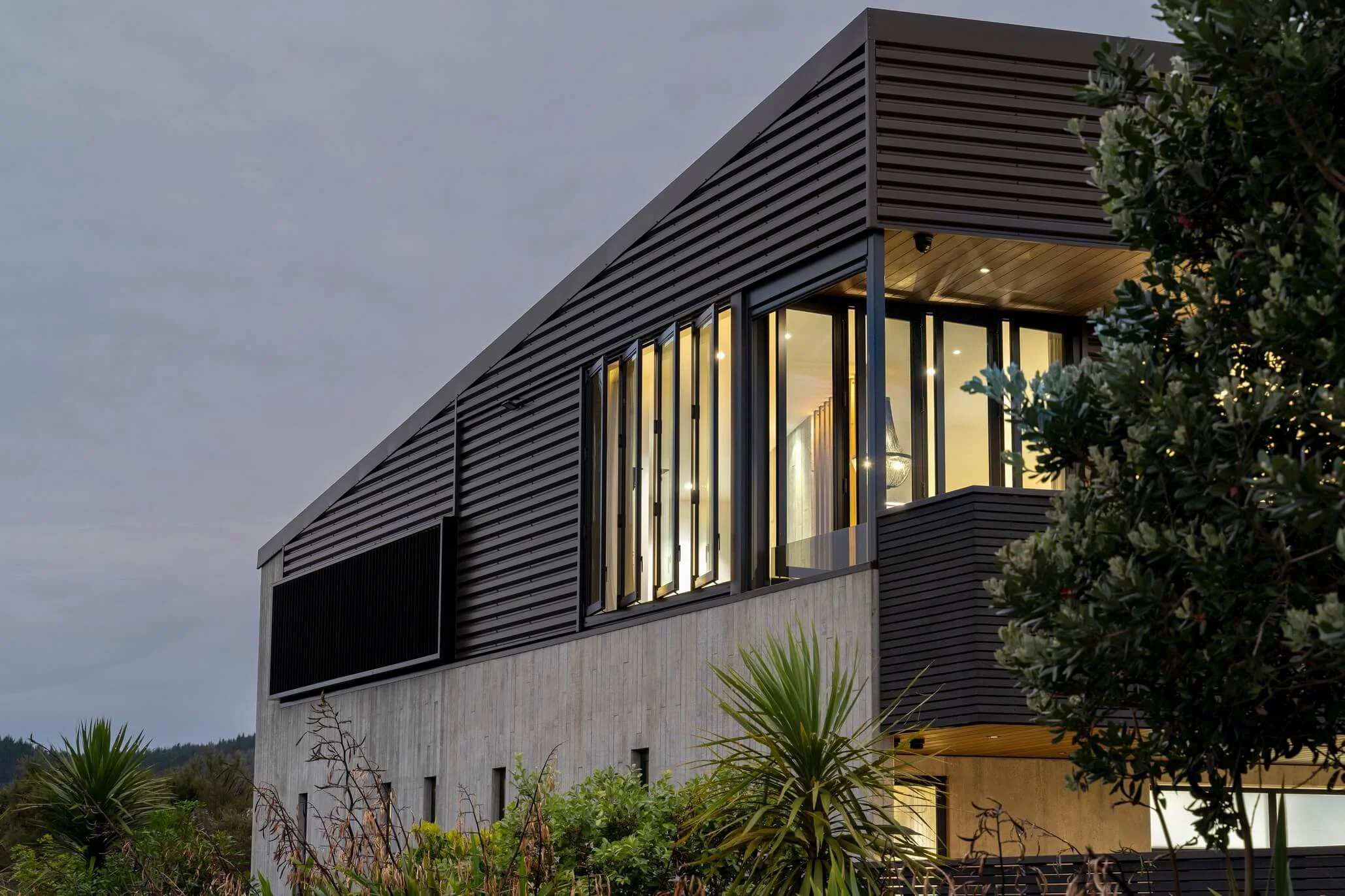 Modern multi-story house with large glass windows and black horizontal siding, surrounded by greenery, under a cloudy sky at dusk.