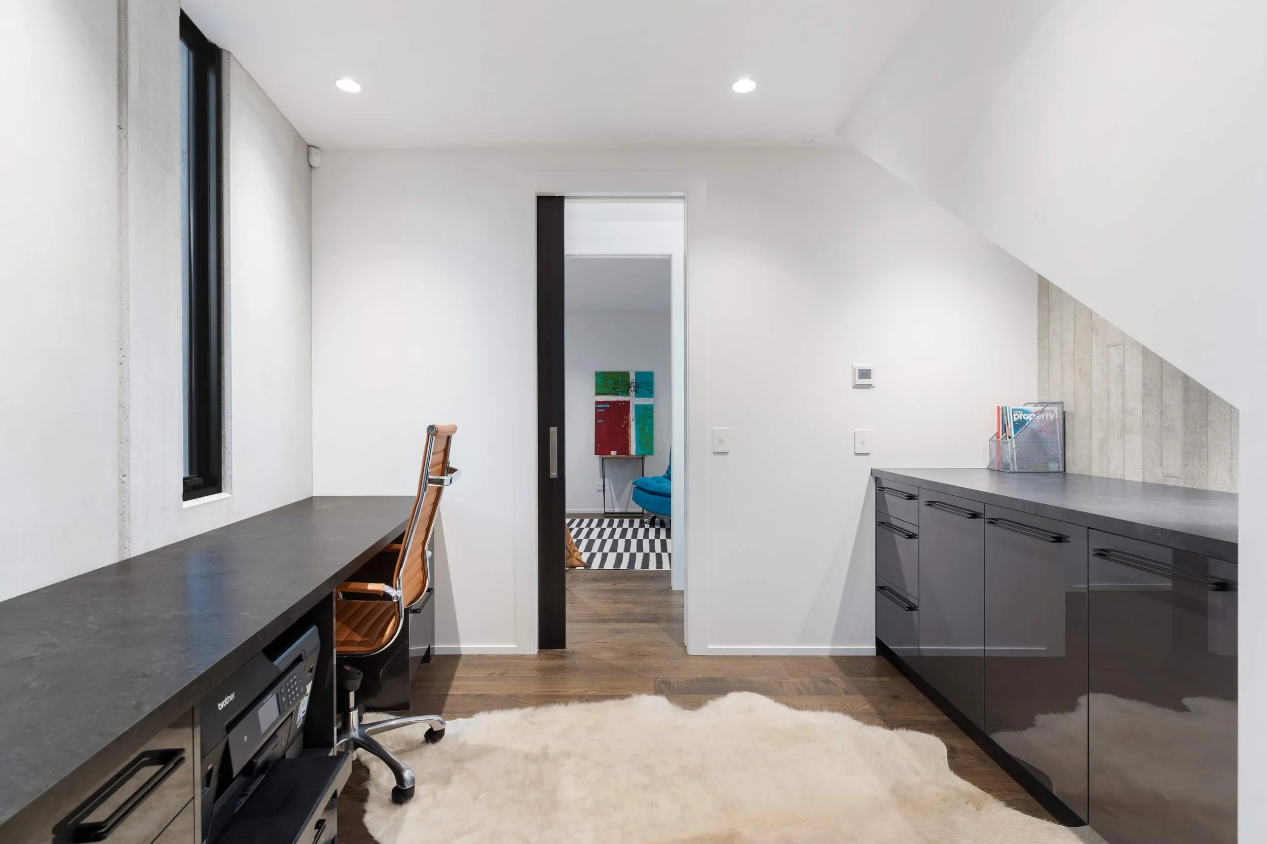 Modern home office with white walls, dark wood flooring, a black countertop on the left with a printer beneath, a peach-colored office chair, a black kitchen countertop on the right, a small wall-mounted thermometer, and a doorway leading to a cozy r