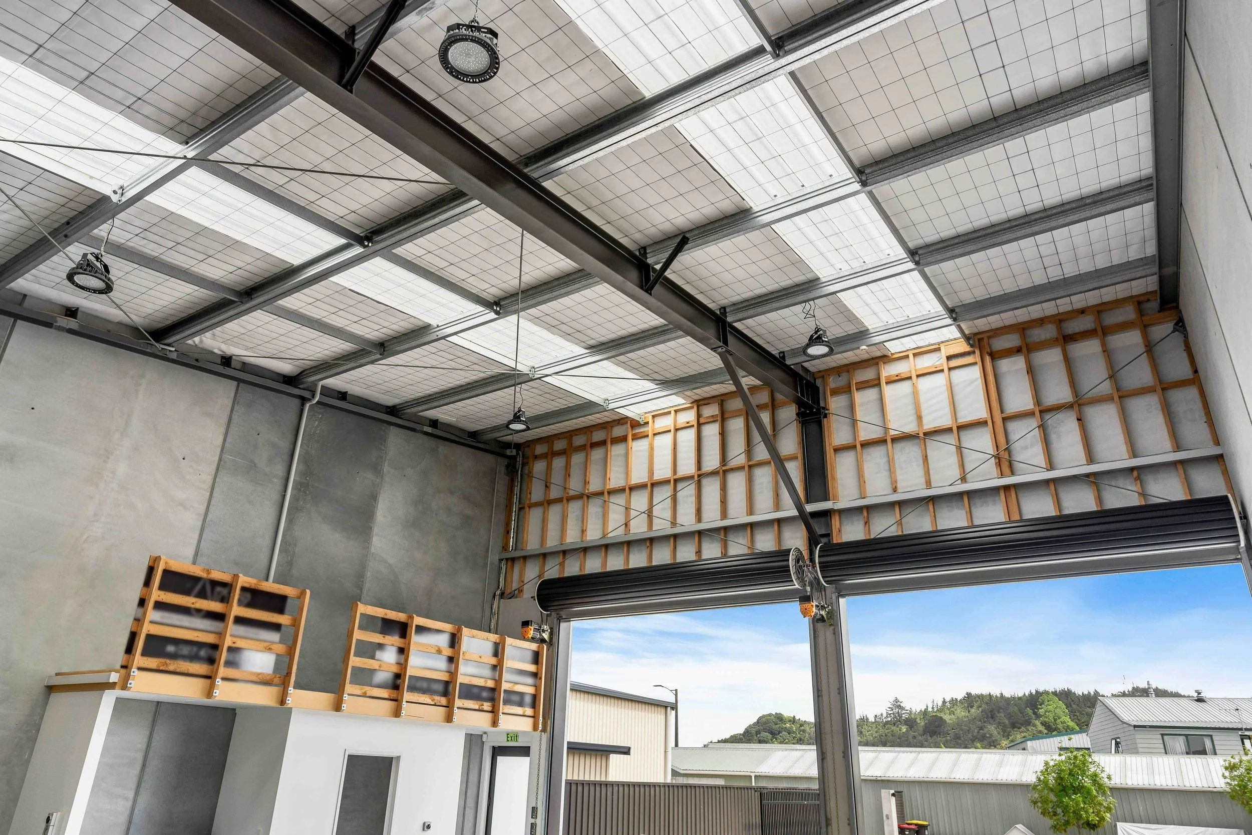 Interior view of a building under construction with steel beams, wooden framing, and ceiling panels, and an open window showing a clear sky and trees outside.