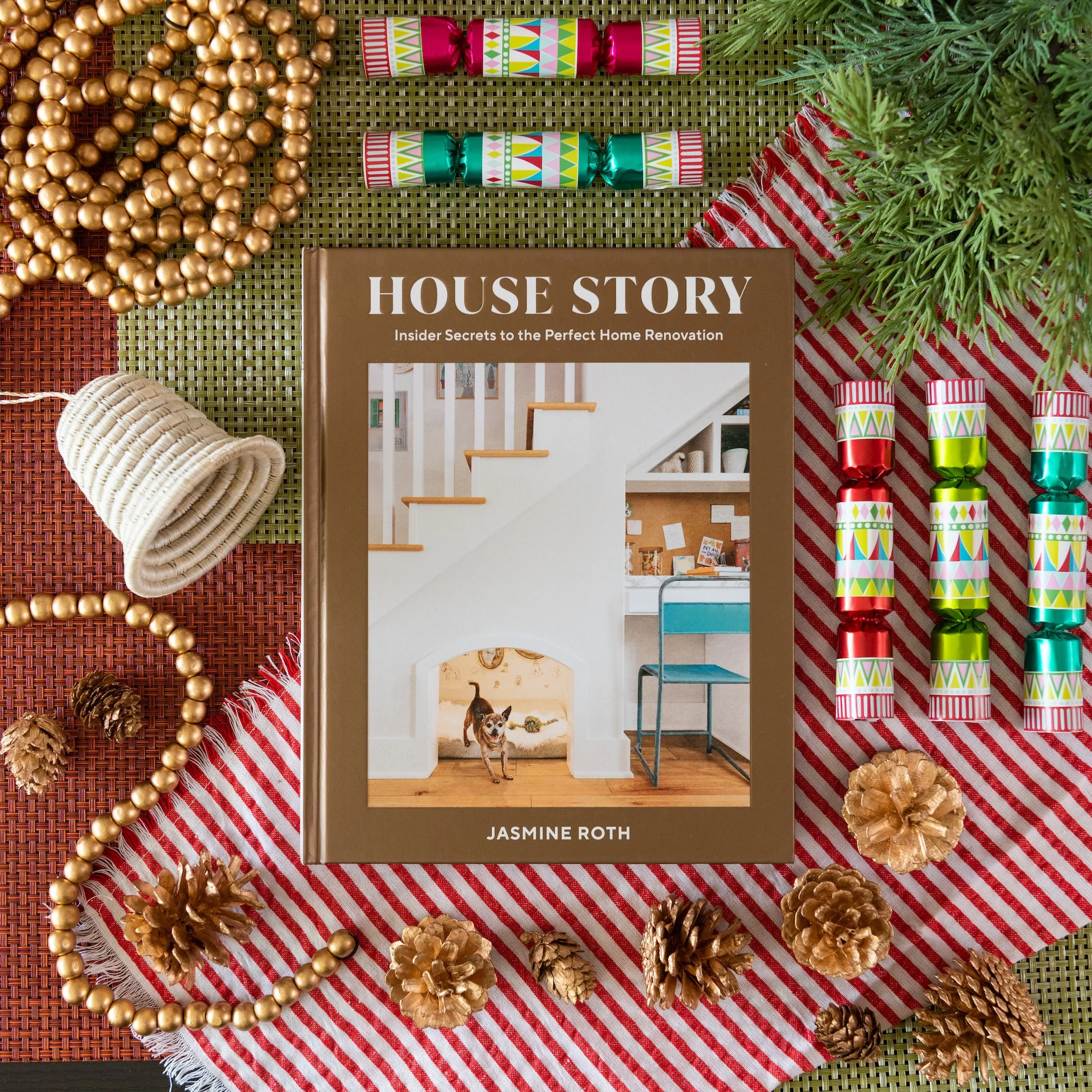 Decorative holiday items arranged on a textured surface including gold beads, pinecones, a holiday-themed book titled 'House Story' by Jasmine Roth, a flat lay styled by Kelli Hart.