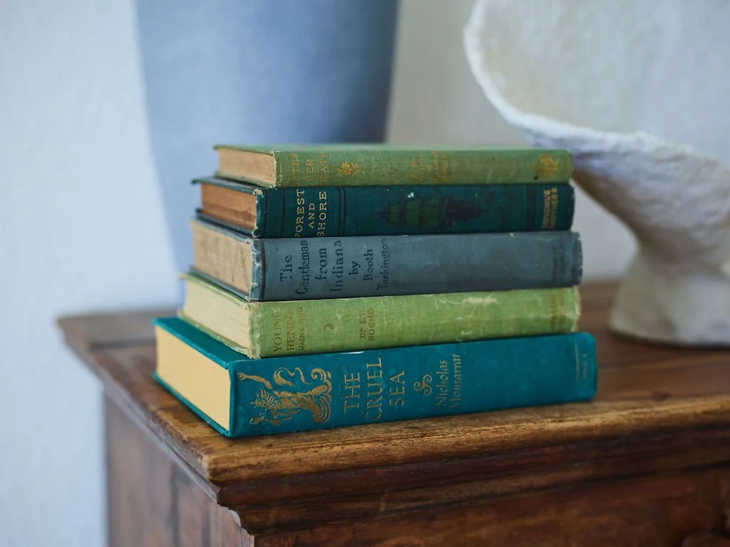 A stack of six vintage hardcover books with worn covers and spines, placed on a wooden surface beside a blue vase and a white decorative object, styled by interior design author and ghostwriter Kelli Hart.