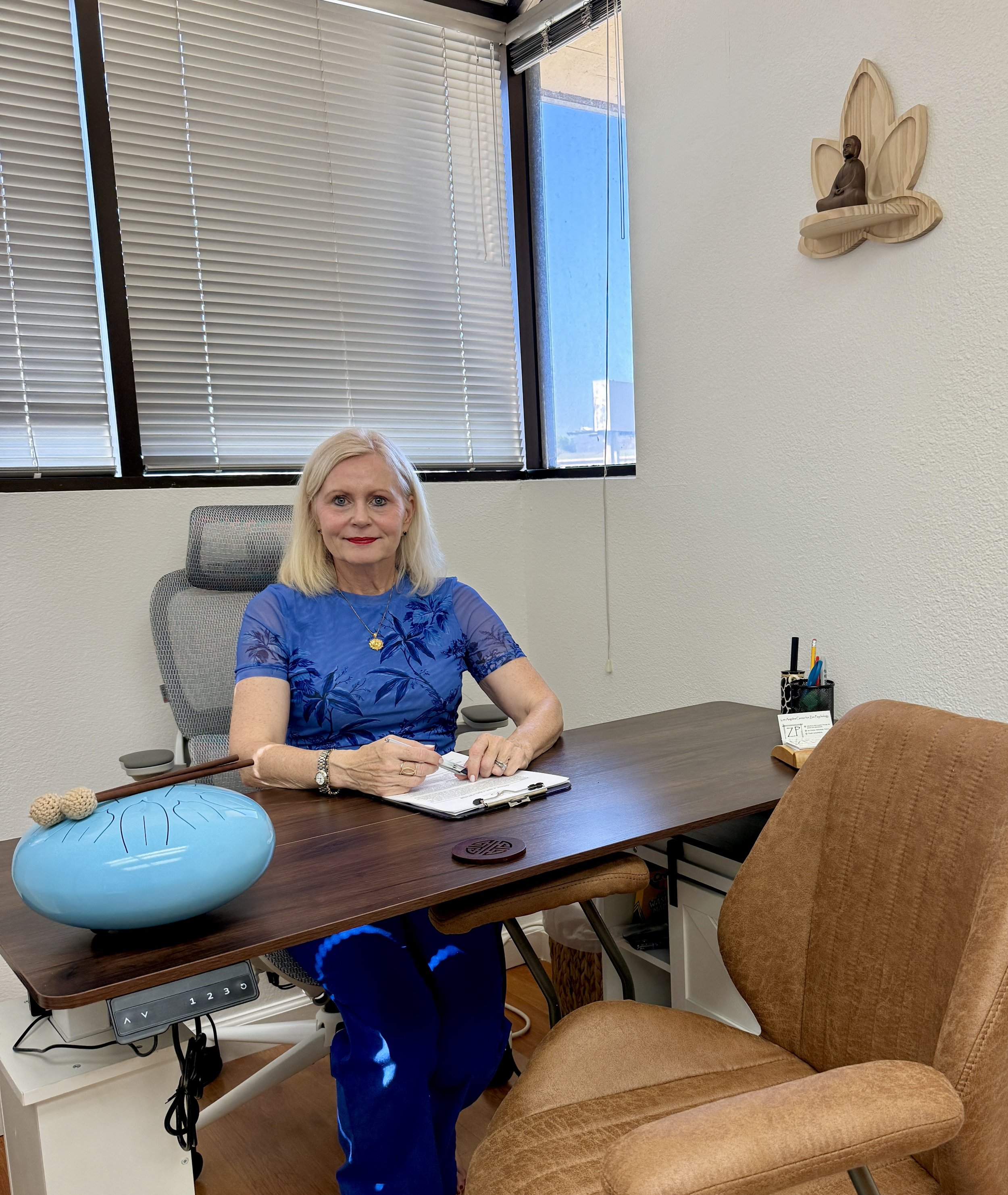 Woman in a blue floral shirt seated at a desk in an office, holding a pen over paperwork, with blinds, a blue decorative bowl, and a Buddha wall carving.