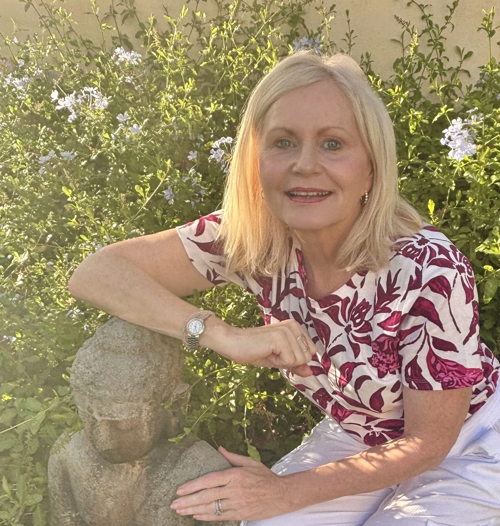 Smiling blonde woman in a floral blouse sitting beside a stone fountain in a sunlit garden