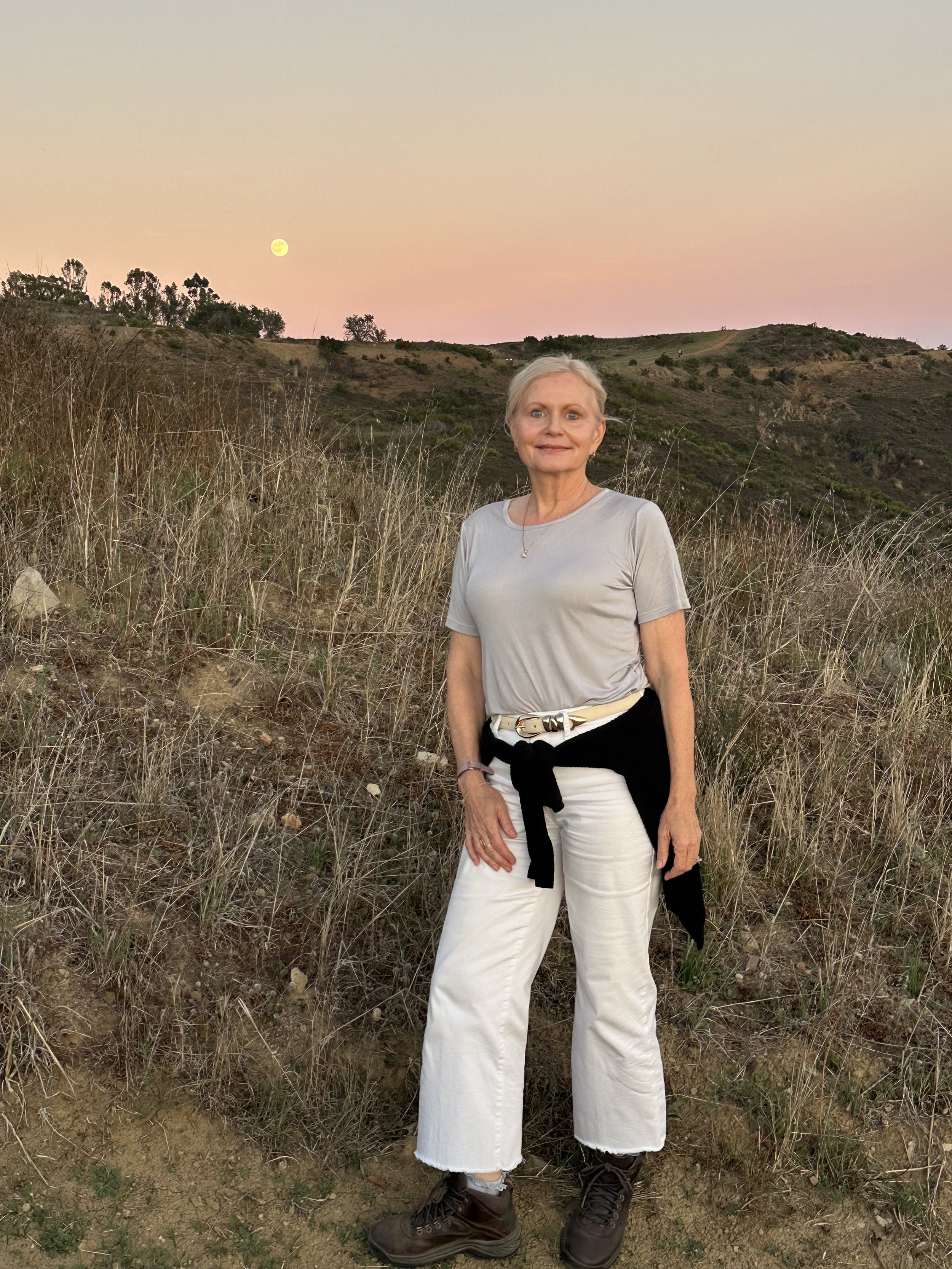 Woman posing in a dry grassy hillside landscape at sunset with the moon visible in a pink sky