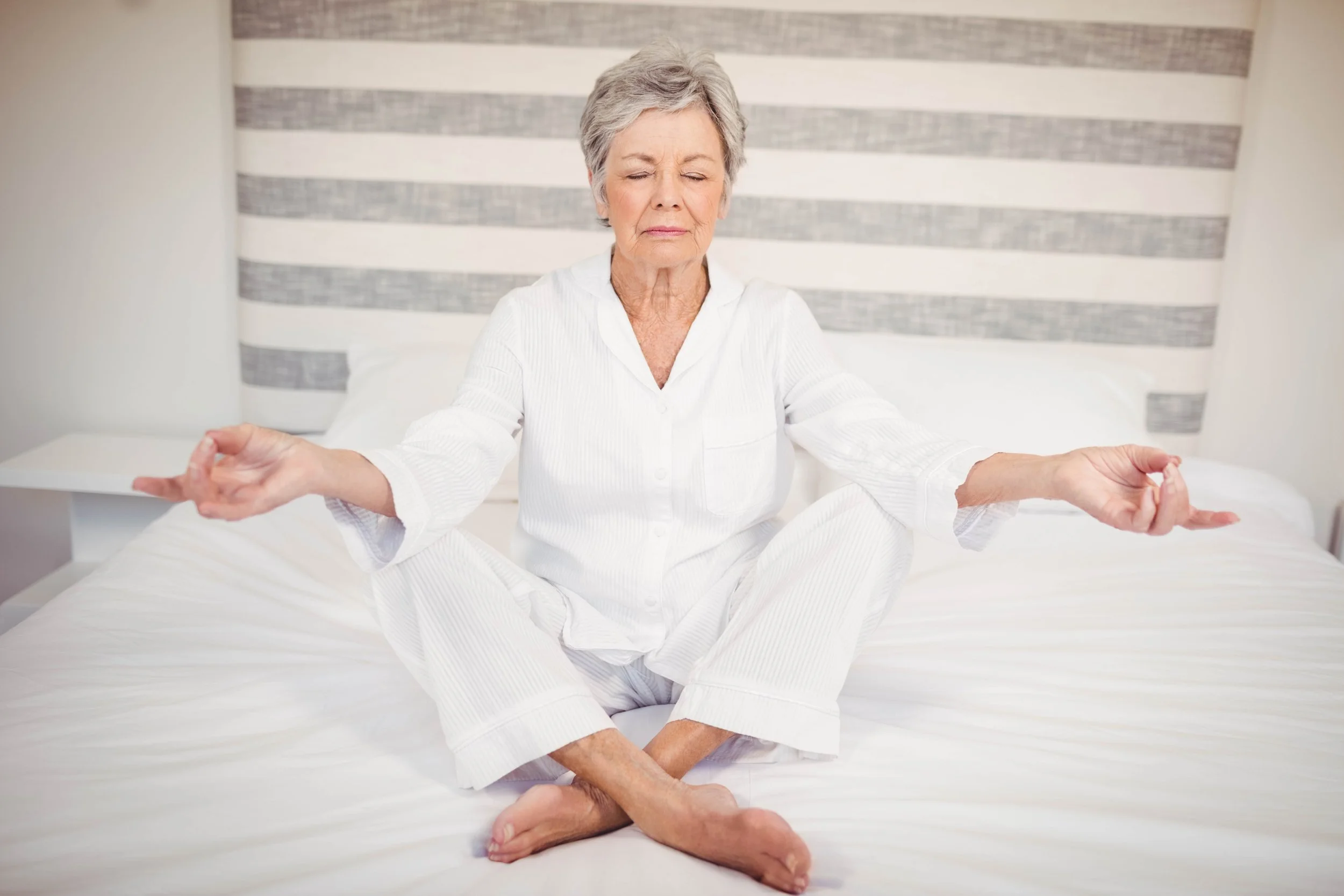 Older woman meditating cross-legged on a bed with eyes closed and hands resting in a yoga pose