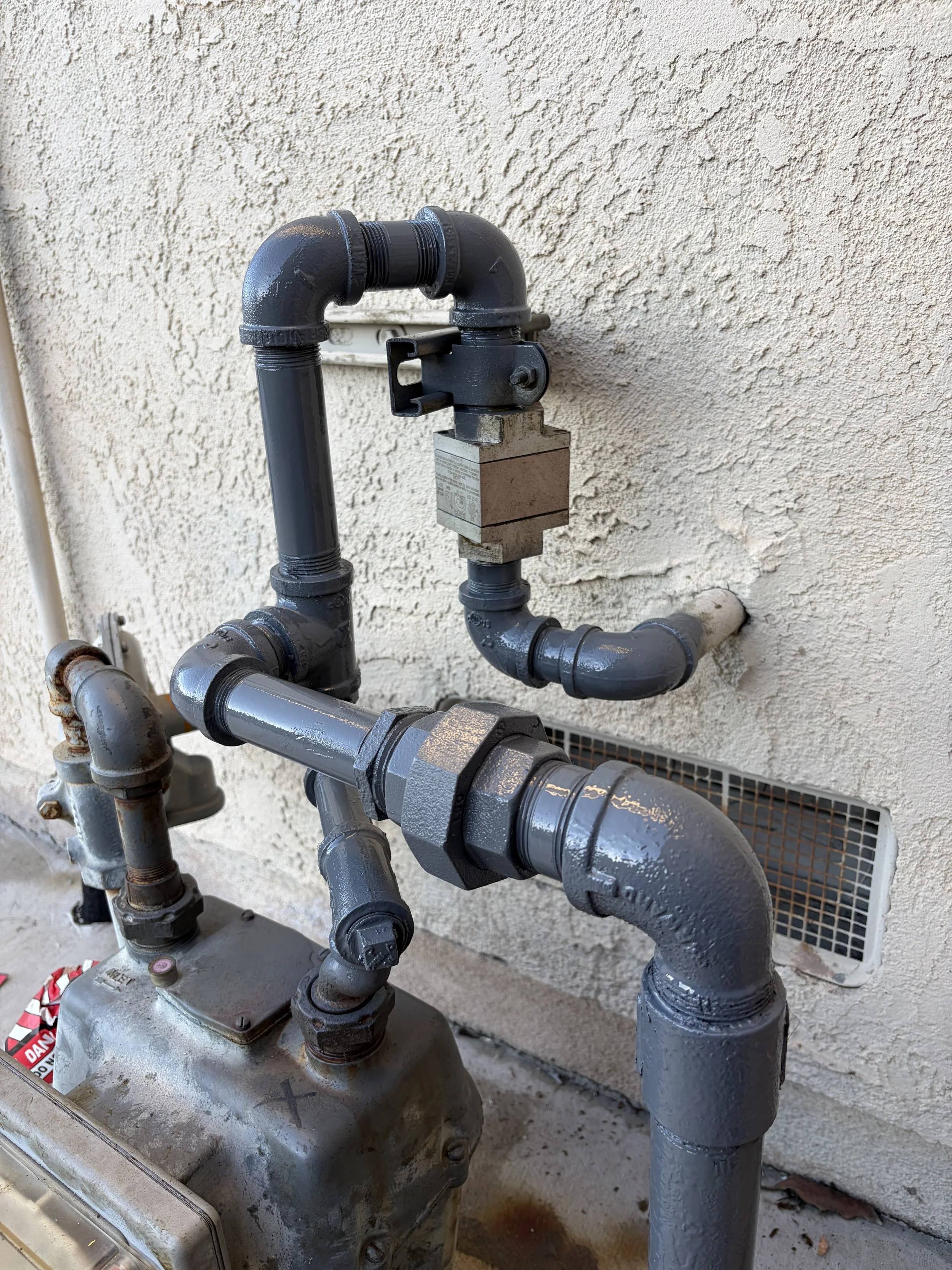 A close-up view of black metallic plumbing pipes mounted on an exterior wall, connected to a gray box or water meter, with a ventilation grate in the background.