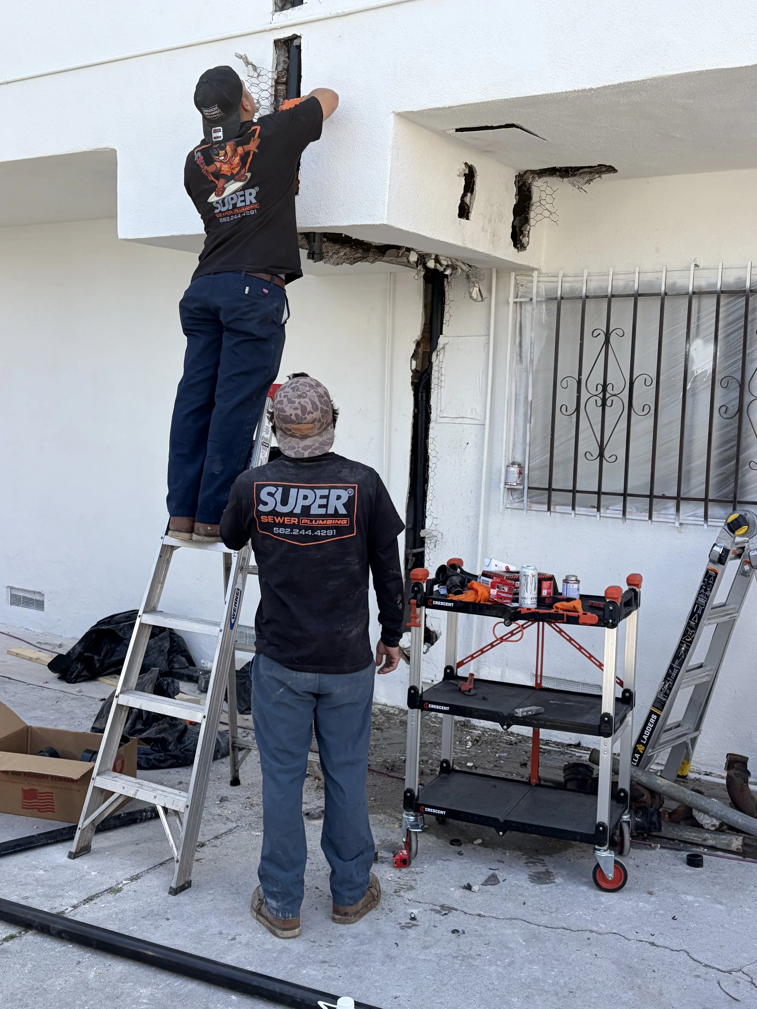 Two workers repairing a hole in the exterior wall of a building using ladders and tools, with work equipment and supplies nearby.