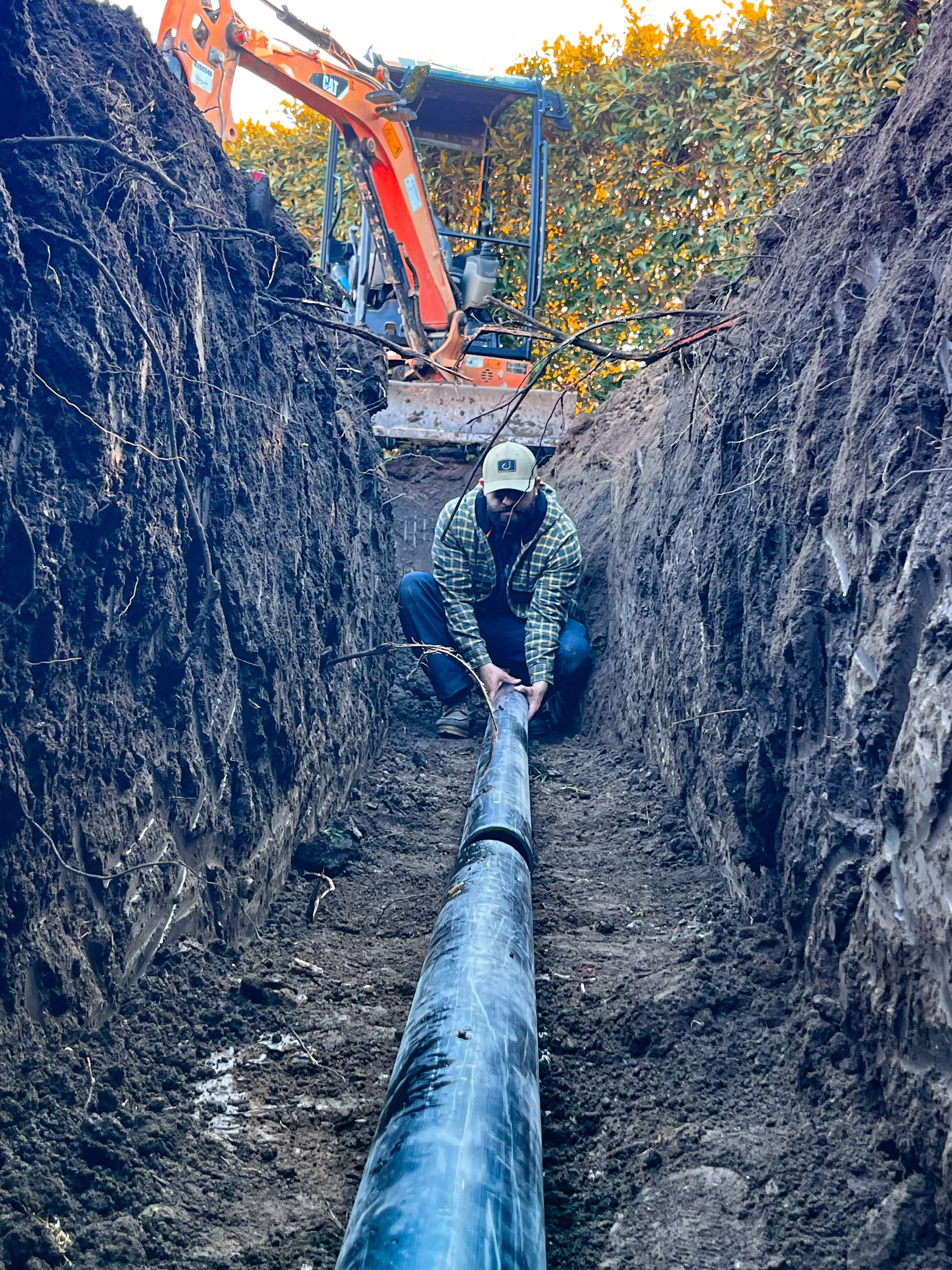 A worker installing a large underground pipe in a trench with a backhoe digging behind him, surrounded by dirt and soil.