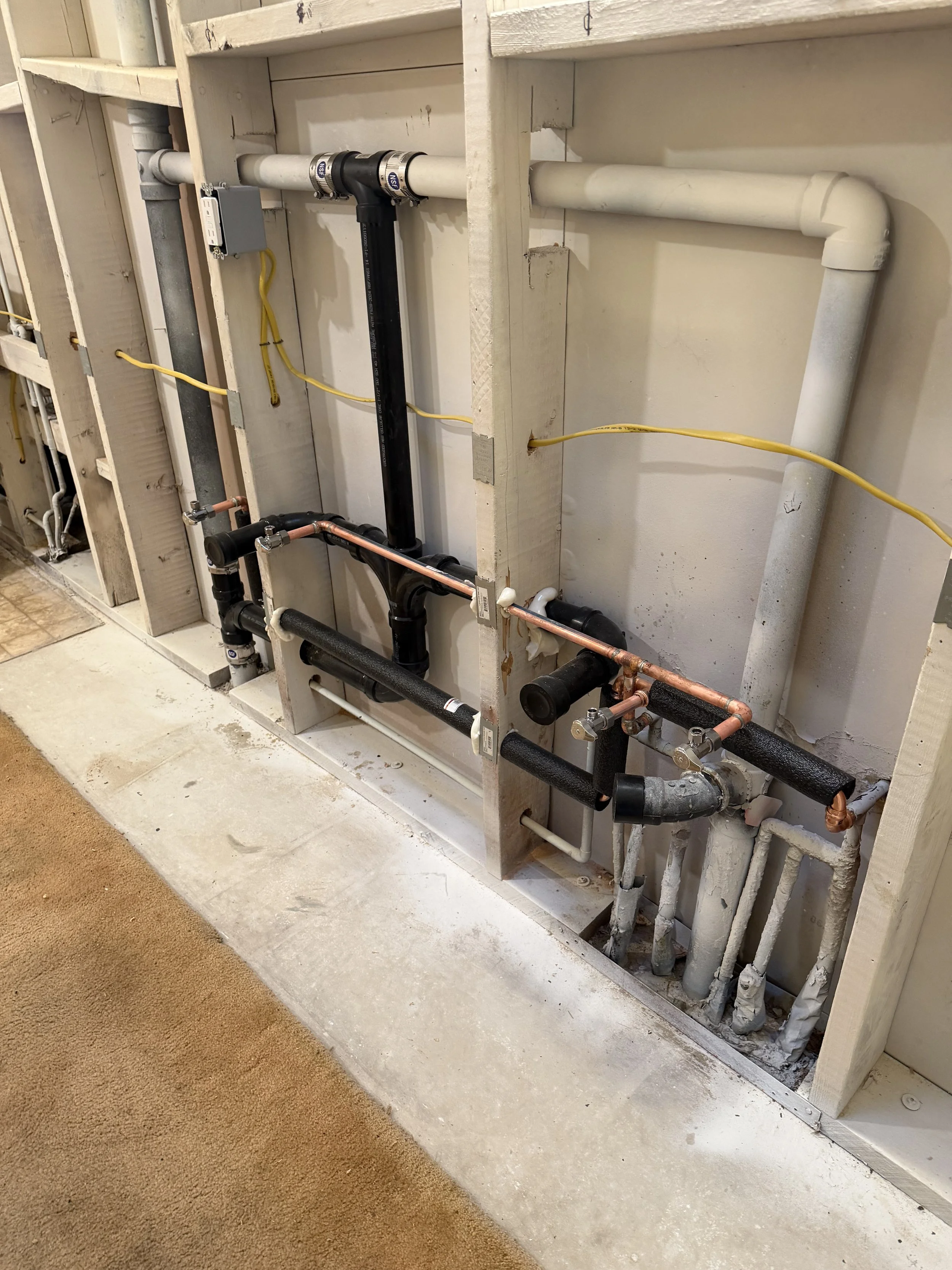 View of plumbing pipes and fittings in a wall cavity, including PVC, copper, and black PEX pipes, with some electrical wiring, in a construction or renovation site.
