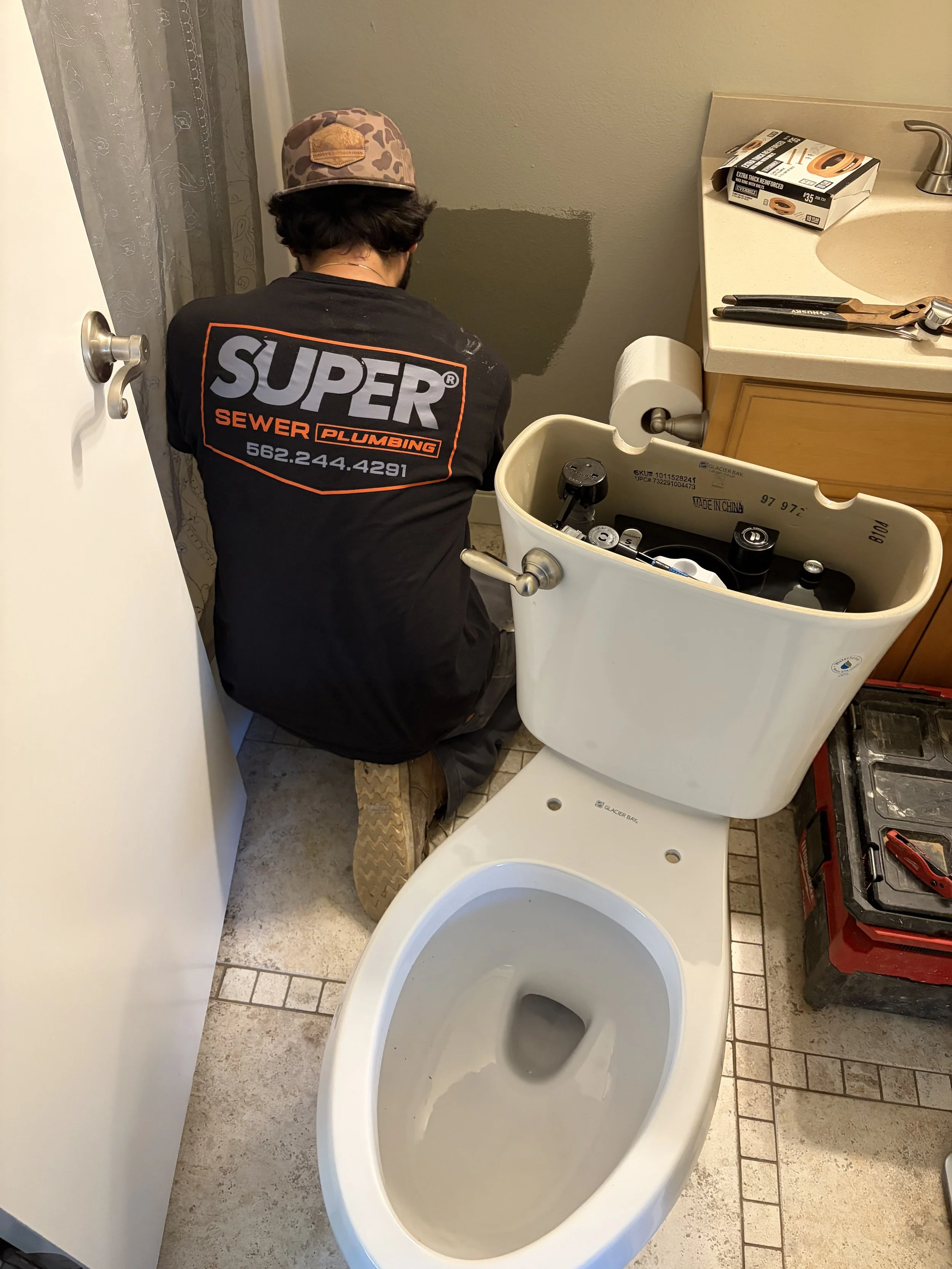 A person kneeling next to a toilet with its tank lid off, working on plumbing. The person is wearing a black shirt with 'SUPER' and 'SEWER PLUMBING' on the back. There are tools and materials on the bathroom counter nearby, including a box of plumbing supplies and a wrench. The bathroom has tiled flooring and a partially painted wall.