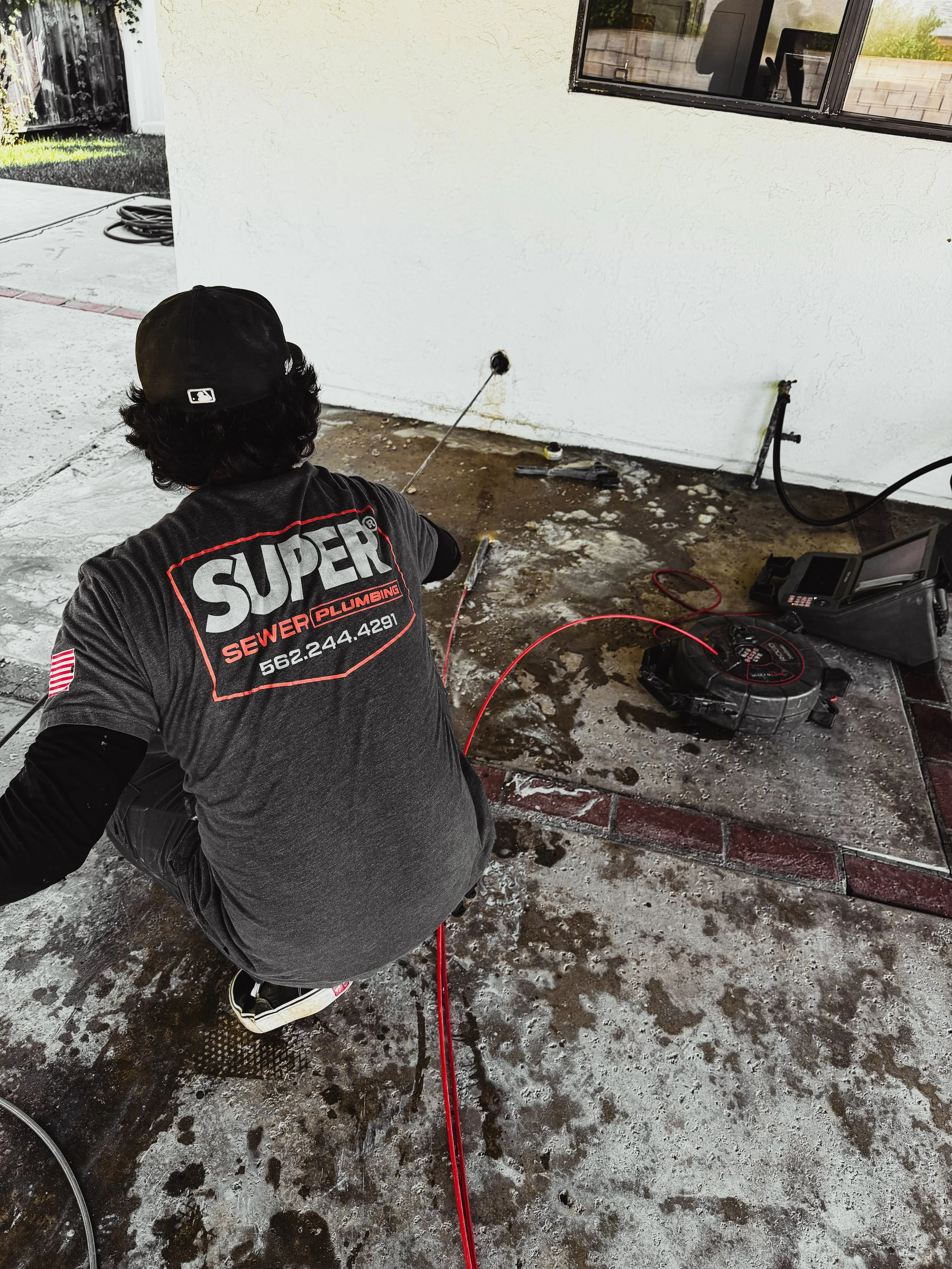 A worker crouching on a concrete patio, using a power tool to drill a hole in the wall of a house. The worker is wearing a black baseball cap and a black T-shirt with a company logo.
