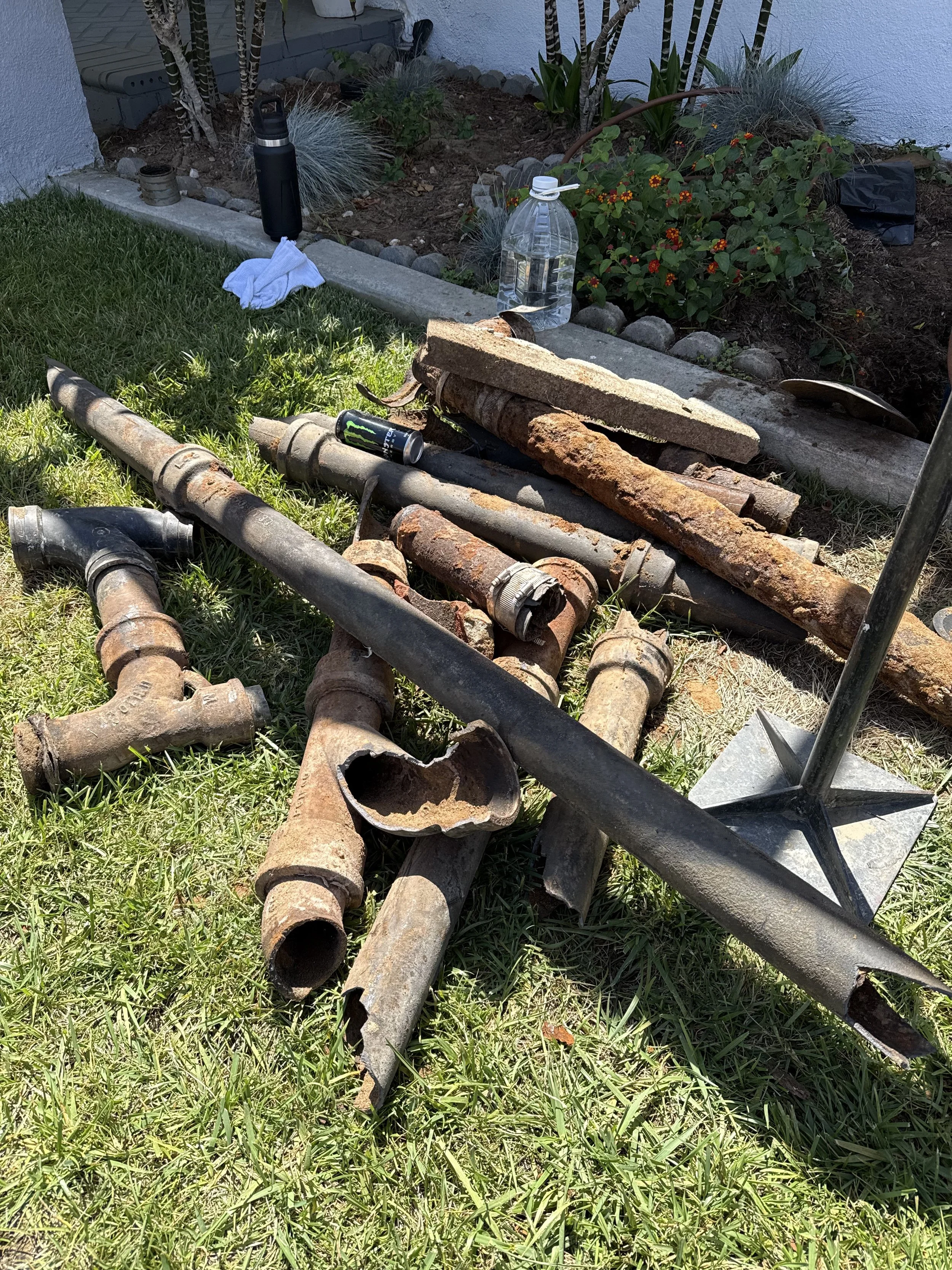 A yard with various rusted metal pipes and pipes fittings, a water bottle, a black water container, and gardening tools, with a flower bed and green grass.