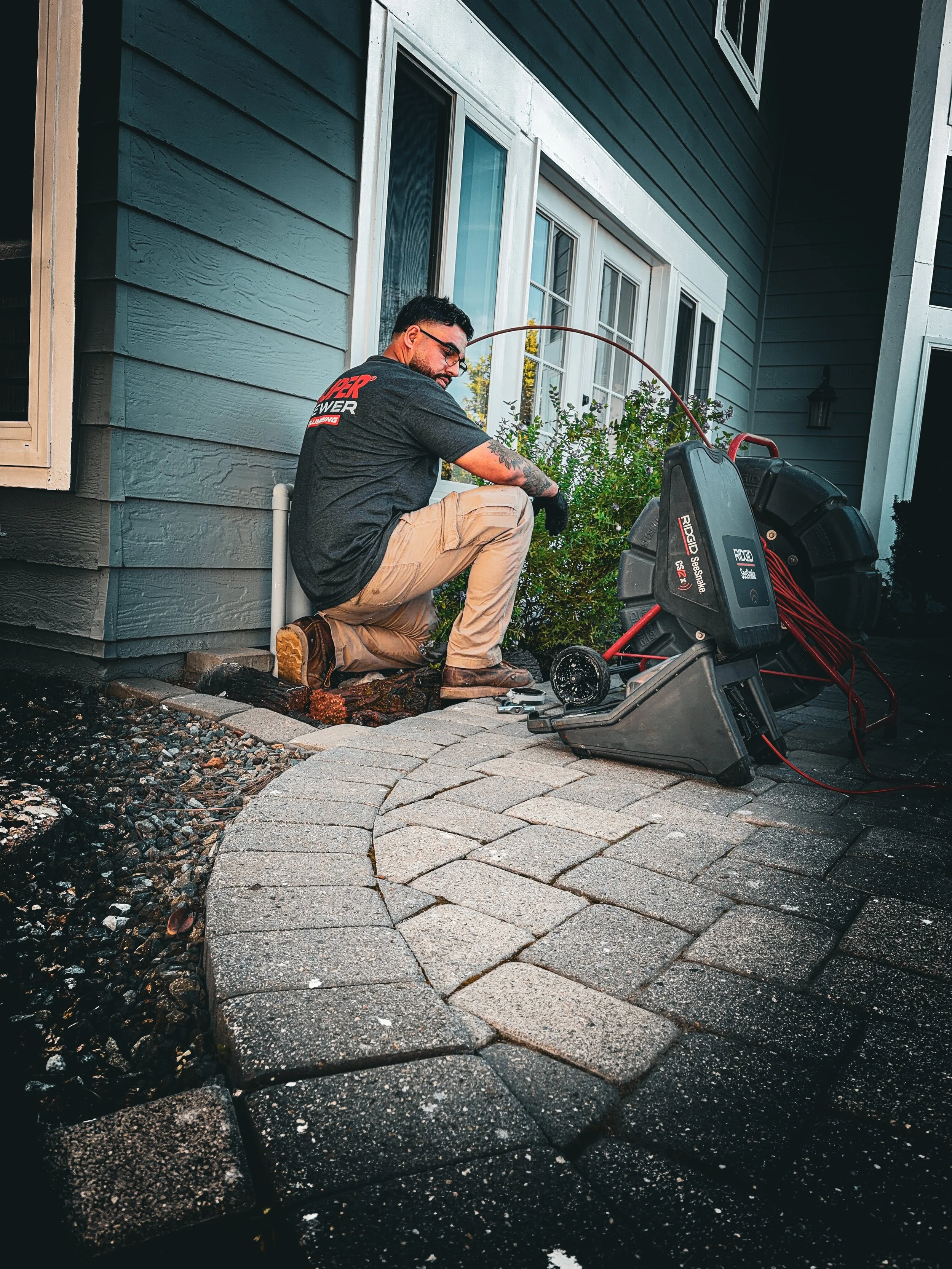 A man kneeling next to a new stone paver walkway outside a house, working with a power tool amid landscaping materials.