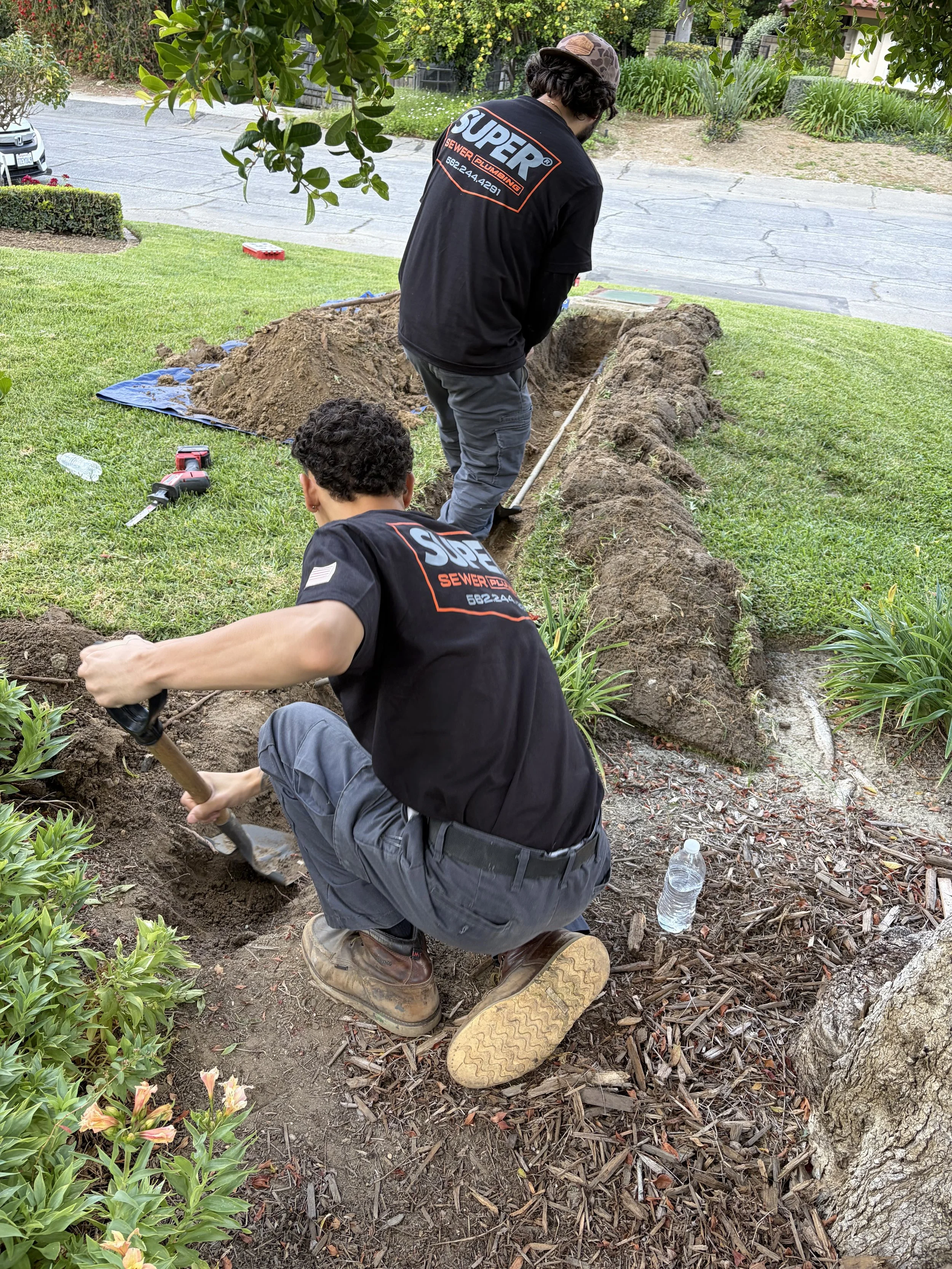 Two workers in black shirts labeled 'SUPER Sewer Plumbing' are digging a trench in a front yard. One is kneeling with a shovel, and the other is standing, both focused on the excavation. Gardening tools, a water bottle, and a water hose are nearby, with a garden and sidewalk in the background.