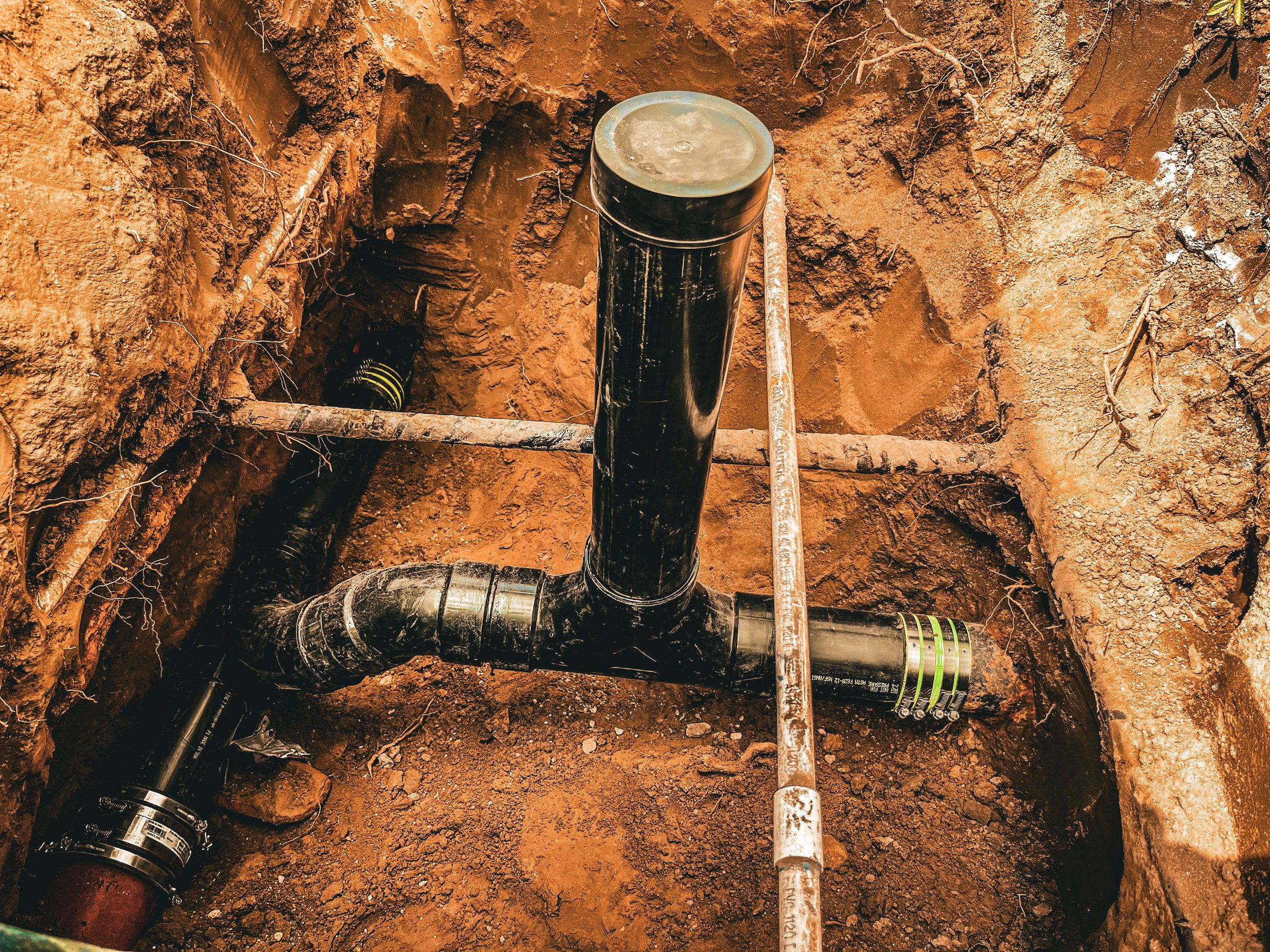 An underground pipe system with black pipes, a white pipe, metal fittings, and a section of a yellow and black pipe, surrounded by exposed dirt and roots.