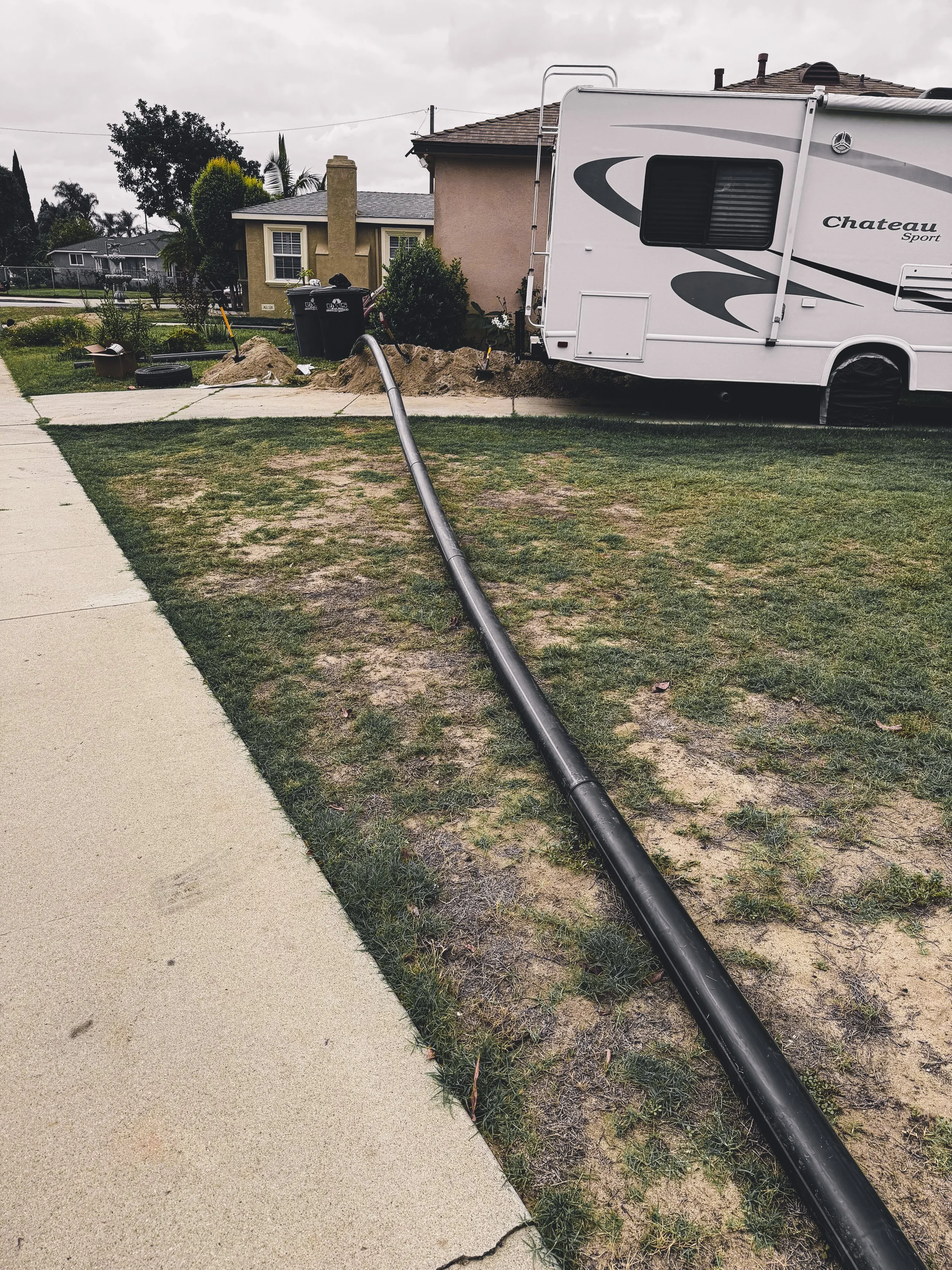 A residential street with a white trailer parked on the grass near the sidewalk. There are construction tools and excavated dirt around, indicating ongoing work.